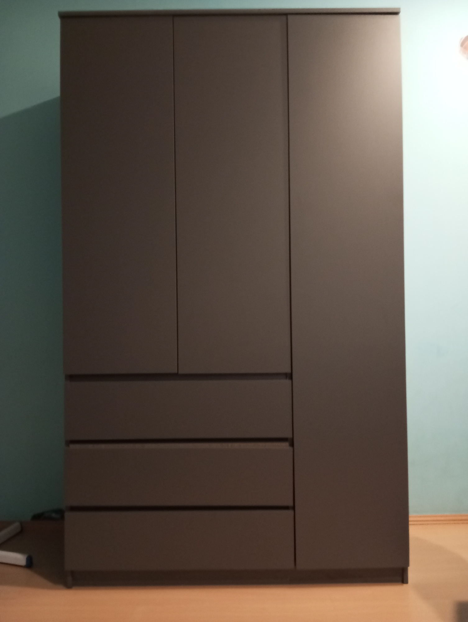 The image shows a large, dark brown wardrobe with four doors, three of which are on the left side and one on the right. Below the left doors are three drawers. The wardrobe is situated in a room with light blue walls and a wooden floor. A light fixture is visible on the right wall, casting a glow. The image appears to be taken from a slightly low angle, looking up at the wardrobe. No people or animals are present. The scene is indoors, in what appears to be a residential setting. The specific location in Verkhnyaya Pyshma, Russia, cannot be determined from the visual information provided in the image.
