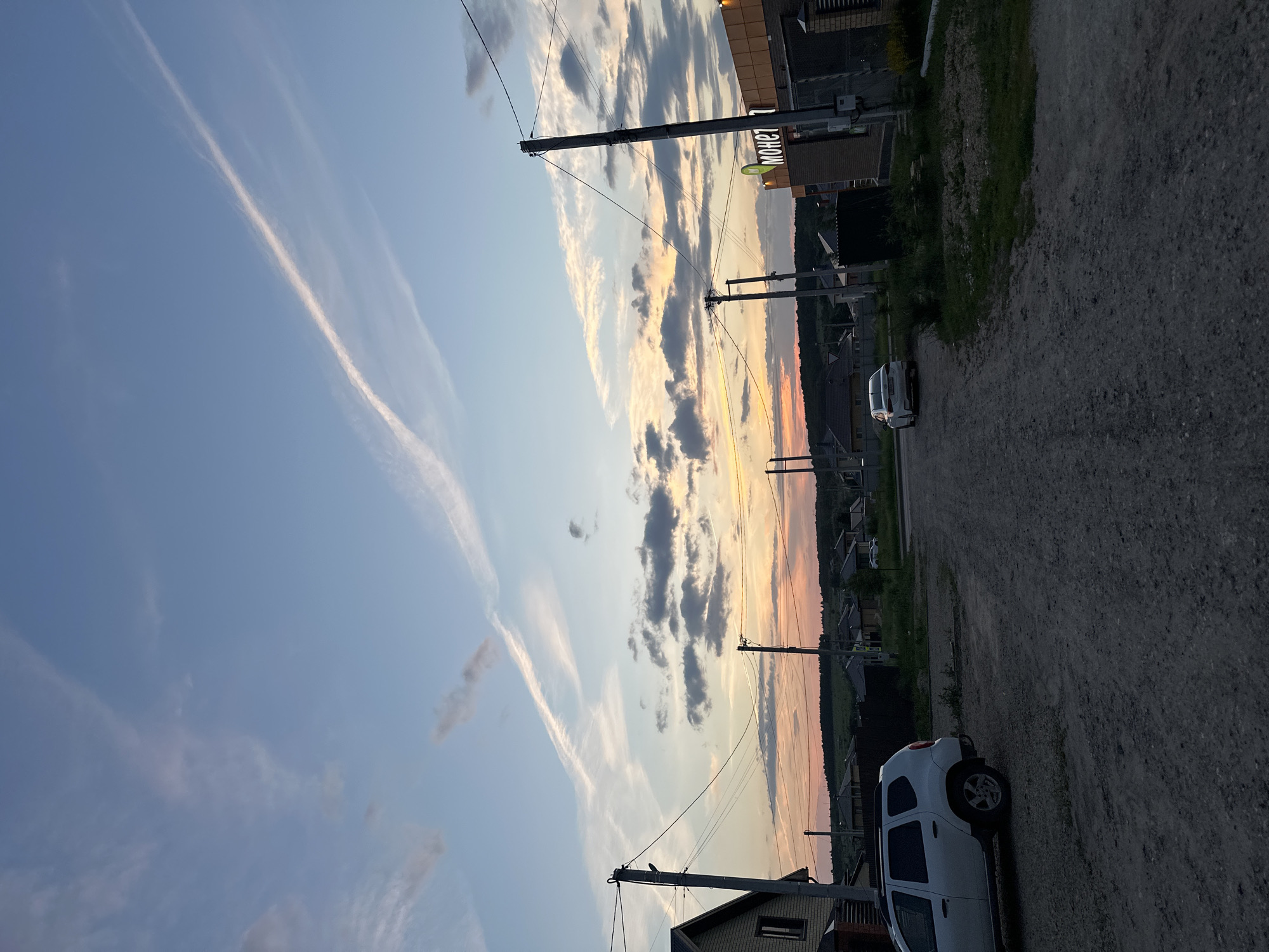 The image shows a gravel road in a rural or suburban area, likely the city of Estachi, Russia, during twilight. The sky is a pale blue with wispy clouds, some tinged with pink and orange from the setting sun. There are power lines crisscrossing the scene, supported by wooden poles. On the left, a white SUV is parked on the side of the road, with a portion of a house visible behind it. Further down the road, a white sedan is driving away from the camera. On the right side of the road, a building with a sign reading "МОНЕТ" is visible. The landscape beyond the road consists of low-lying hills dotted with houses and trees. The overall impression is of a quiet evening in a residential area.