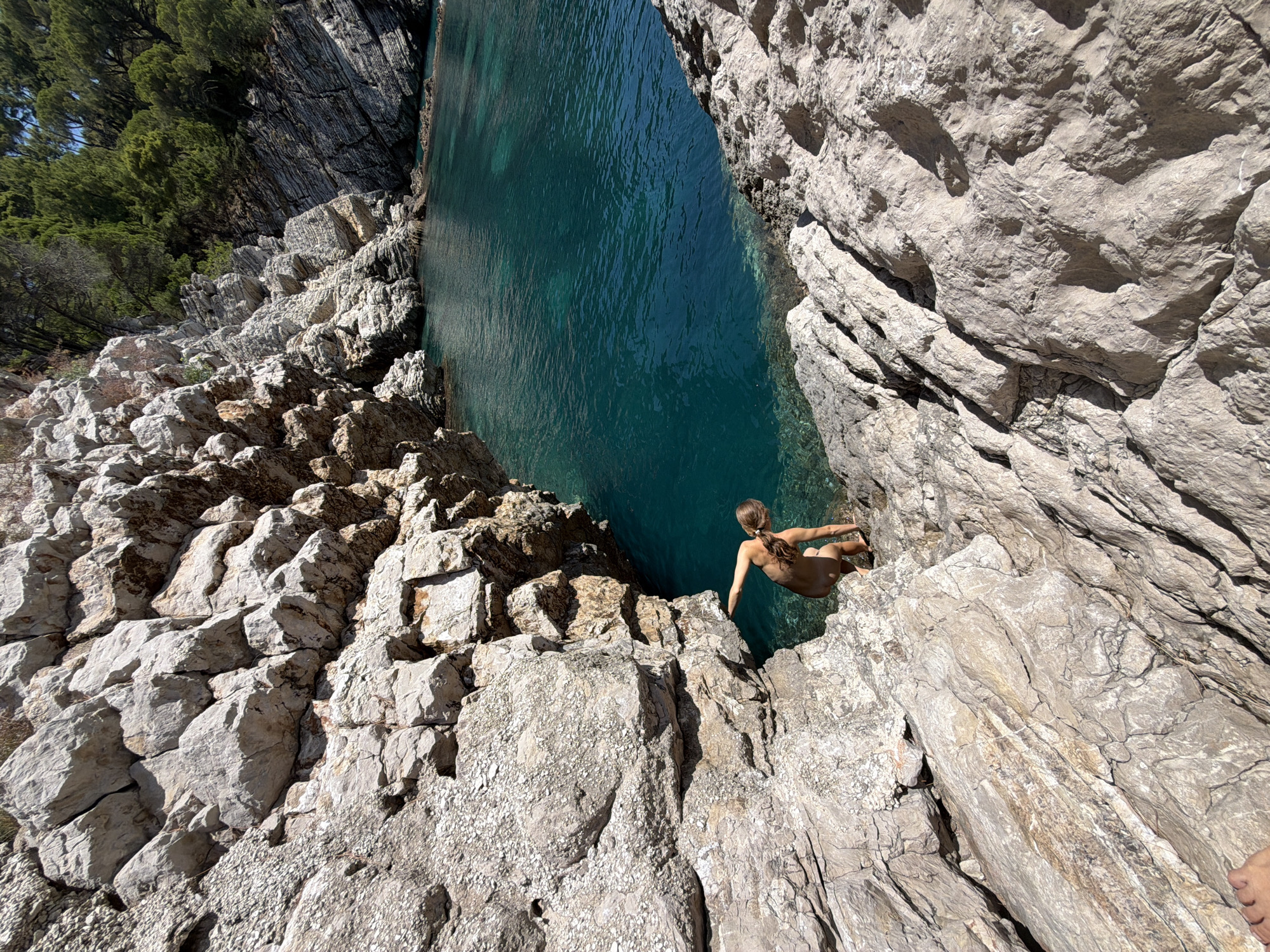 An adult female, with blonde hair pulled back in a ponytail, wearing a dark colored bikini bottom, is captured in a full body shot, emerging from the turquoise waters of a cove. She is positioned on the right side of the frame, partially obscured by rocky outcrops. The rocks are light gray and textured, with some showing signs of weathering. The cove is surrounded by steep, rocky cliffs, with vegetation visible on the higher slopes. The water is clear and reflects the bright sunlight. The scene is likely an outdoor, natural setting, possibly a secluded beach or a rocky inlet. The specific location is Mirište, Montenegro.