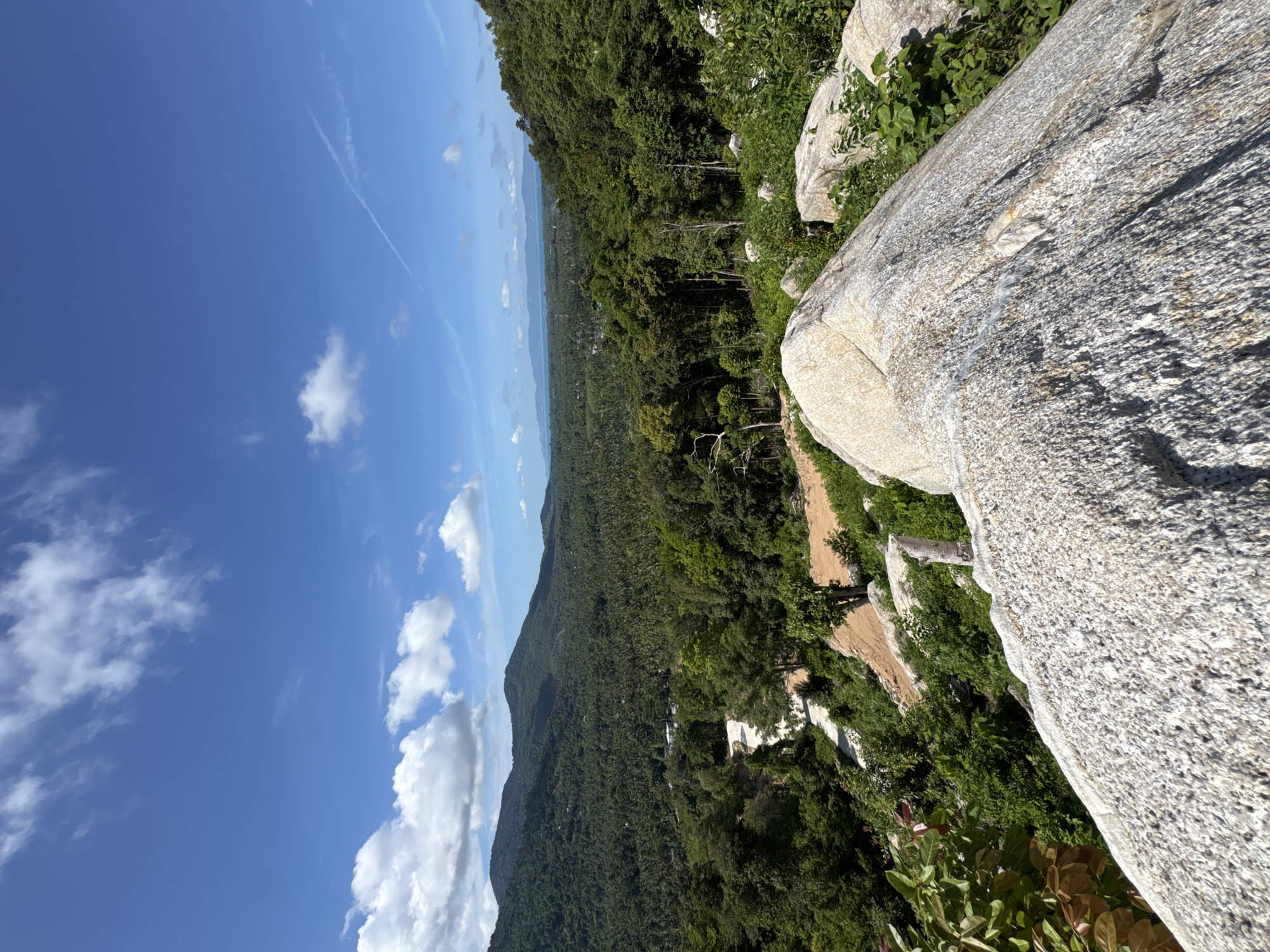 The image is a wide shot of a lush green landscape under a bright blue sky with scattered clouds, likely taken from a high vantage point on Koh Phangan, Thailand. In the foreground, large, rough-textured granite boulders dominate the lower portion of the frame, with smaller rocks and green foliage surrounding them.
Beyond the boulders, the scene opens up to a vast expanse of dense, tropical forest that blankets rolling hills and extends towards the horizon. A winding, light-colored road or path cuts through the vegetation, descending into the valley below. In the distance, the faint outline of islands or landmasses can be seen across a calm body of water, meeting the horizon under a clear sky with a few wispy contrails. The overall impression is one of natural beauty and tranquility.