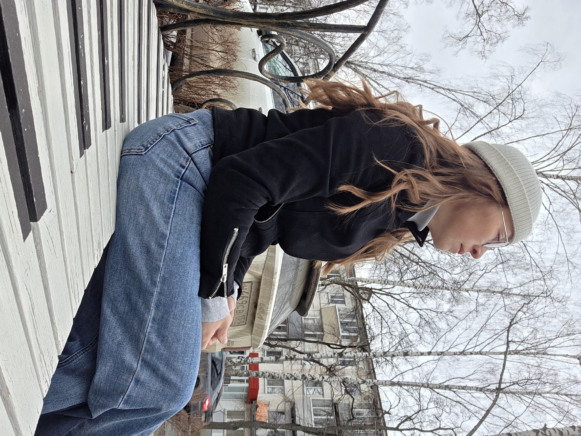 A young woman is standing outside in an urban environment, likely in Chaikovsky, Russia. She is wearing a black jacket, blue jeans, and a white beanie, and she has long, wavy reddish-brown hair. She's leaning against a black and white striped object that resembles a keyboard. The background includes bare trees, a building with windows, and parked cars. The sky is overcast. A wrought iron fence is visible behind her.