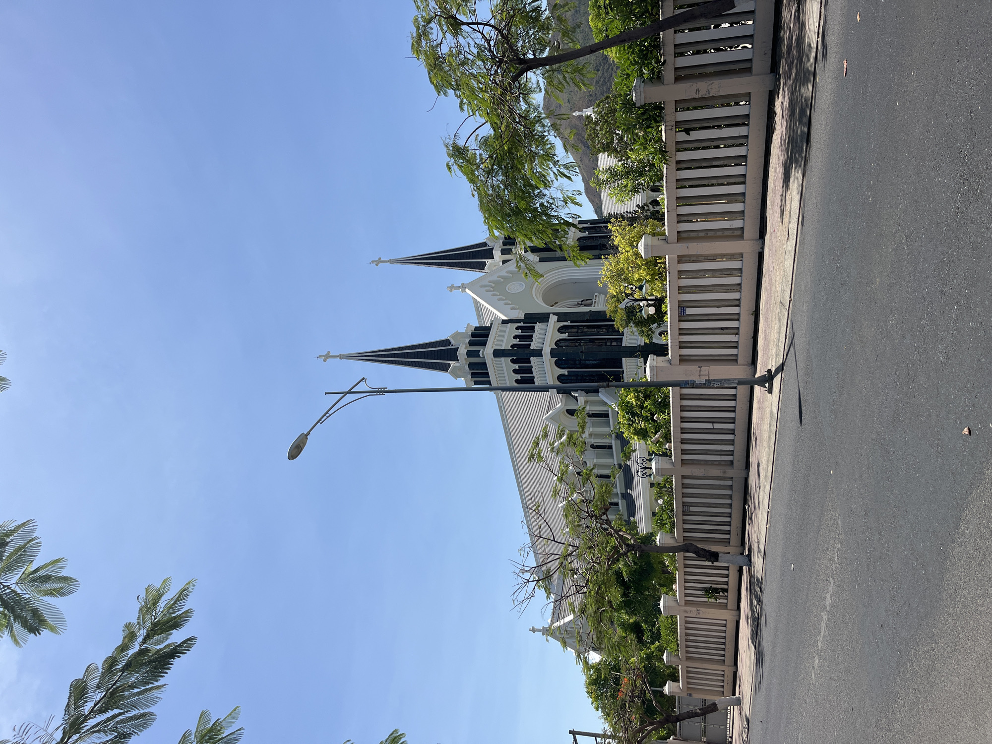 The image shows a street view in Bac Nha Trang Ward, Vietnam, featuring a church as the primary subject. The church is white with black spires and located behind a light-colored fence with greenery growing behind it. A street and sidewalk border the fence. A street lamp is visible near the church. The sky is clear and blue. The angle of the image suggests it was taken from a standing position on the street. There are trees with green foliage on either side of the church, and a mountain can be seen in the background.