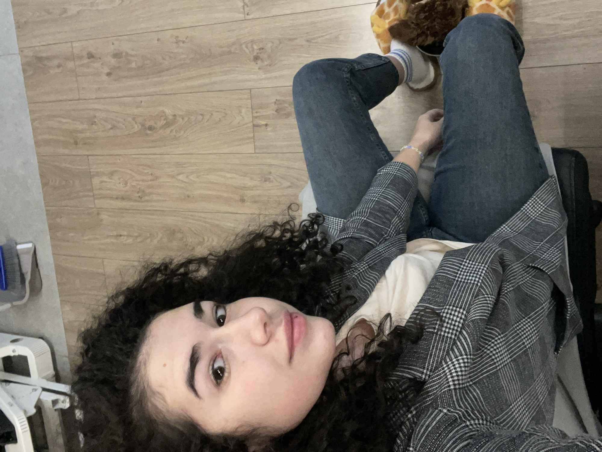 The media file presents a close-up, high-angle shot of a young woman in an indoor setting, likely in Sofia, Bulgaria. She has dark, voluminous curly hair and brown eyes, looking directly at the camera with a neutral and calm expression. She is wearing a grey and white plaid blazer or jacket over a light-colored top, paired with blue denim jeans. A small, colorful beaded bracelet adorns her left wrist.
Her legs are bent, revealing white socks with blue stripes and what appear to be fluffy, animal-themed slippers, possibly resembling giraffes. She seems to be in a relaxed posture, perhaps lying down or leaning back on the floor. The background is primarily a light-colored wooden floor with visible grain and plank patterns. To the far left, a strip of a lighter, possibly grey, floor or wall is visible, along with a partial view of a light-colored object that has a blue brush-like attachment, suggesting household or office equipment. There is no visible text in the image.