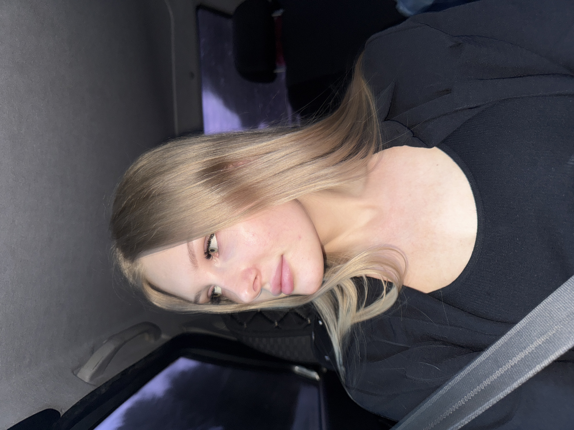 A blonde-haired woman with fair skin is seated in a car. She is wearing a black top and a seatbelt. The interior of the car is visible, including parts of the dashboard and headrest. The location is not possible to be determined based on the image.