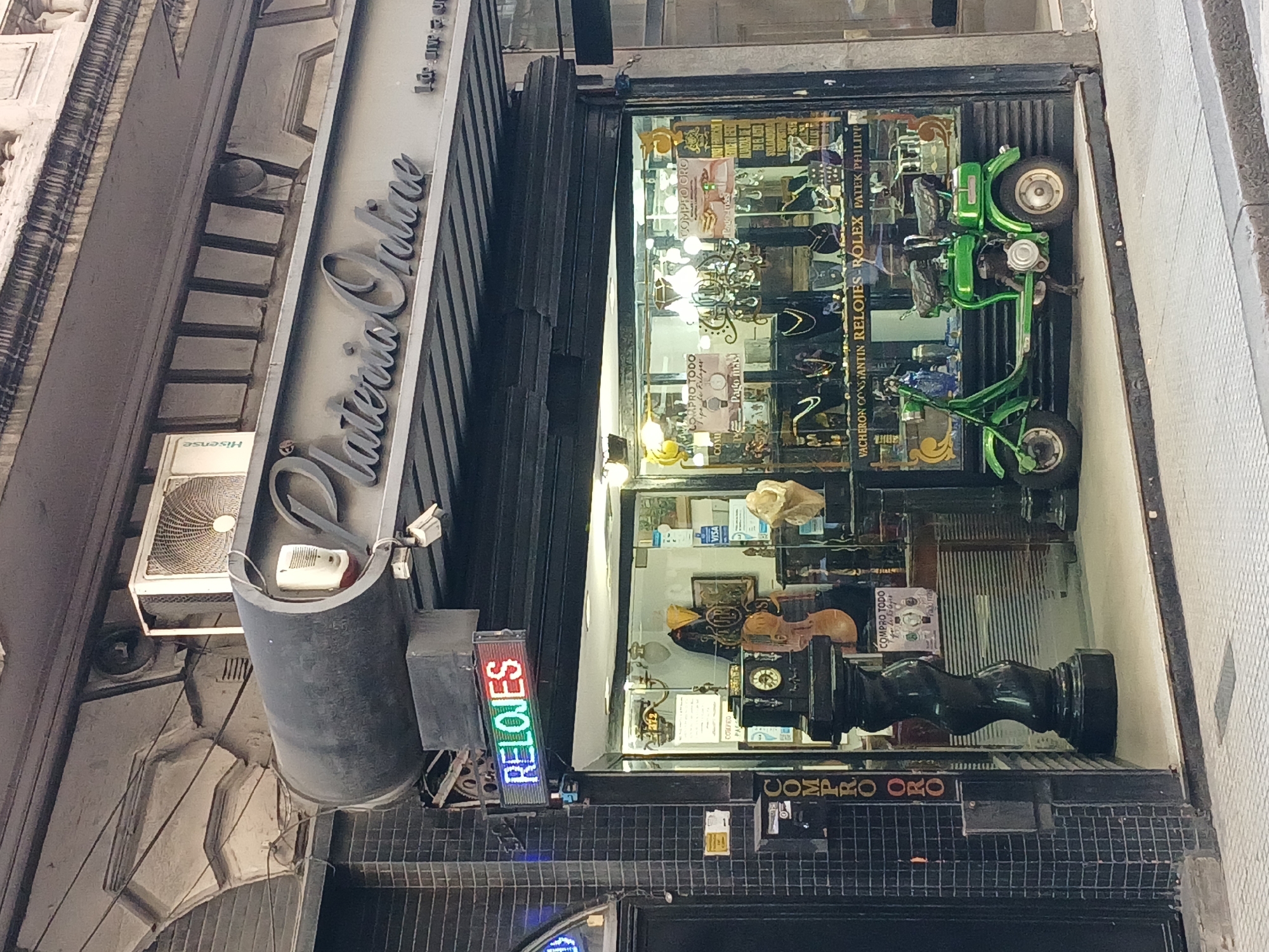 The image is an exterior view of "Plateria Oralice," a jewelry store located in Buenos Aires, Argentina. The store's signage is prominently displayed, along with an illuminated "RELOJES" (watches) sign. The storefront features a glass display window containing various items, including watches (Rolex, Patek Phillippe, Vacheron Constantin) and a green scooter. An air conditioning unit (Hisense) is visible above the entrance. The exterior walls exhibit architectural details with a classical style. "COMPRO ORO" is also visible.