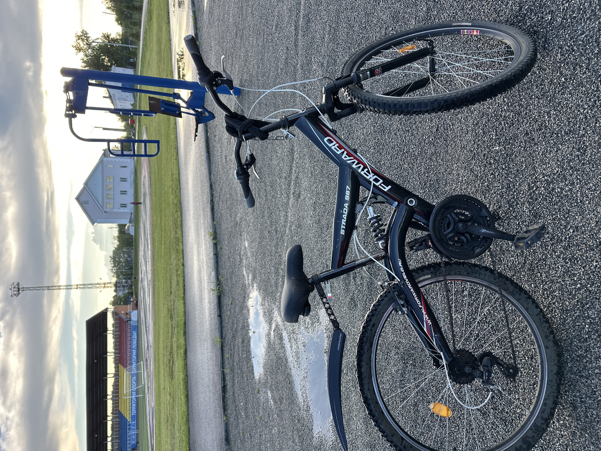 The image displays an outdoor scene in Yuzhnouralsk, Russia, under an overcast sky with filtered daylight. In the foreground, a black "FORWARD STRADA 987" full-suspension mountain bike is parked on an asphalt surface embedded with small gray gravel. The bike features black front and rear fenders, a front disc brake, a standard rim brake on the rear wheel, white cable housing, and orange wheel reflectors. Visible dirt adheres to the tires and fenders. Several puddles on the ground reflect the sky.
In the midground, a blue outdoor exercise machine, resembling a chest press or rowing apparatus, is positioned to the right of the bicycle. Beyond this, a paved track encircles a green grassy field.
The background reveals a sports stadium with covered stands featuring distinct sections of blue, yellow, and red seating. A banner displayed along the stadium wall reads in Cyrillic: "ТЕРРИТОРИЯ СПОРТА - ТЕРРИТОРИЯ ПОБЕДЫ!", which translates to "TERRITORY OF SPORT - TERRITORY OF VICTORY!". A tall metal light tower with a fixture at its apex stands behind the stadium. To the right, a light blue and white two-story building with multiple windows is visible, along with a smaller, lighter-colored building further beyond. Scattered trees line the distant horizon. No identifiable individuals are clearly discernible within the frame.