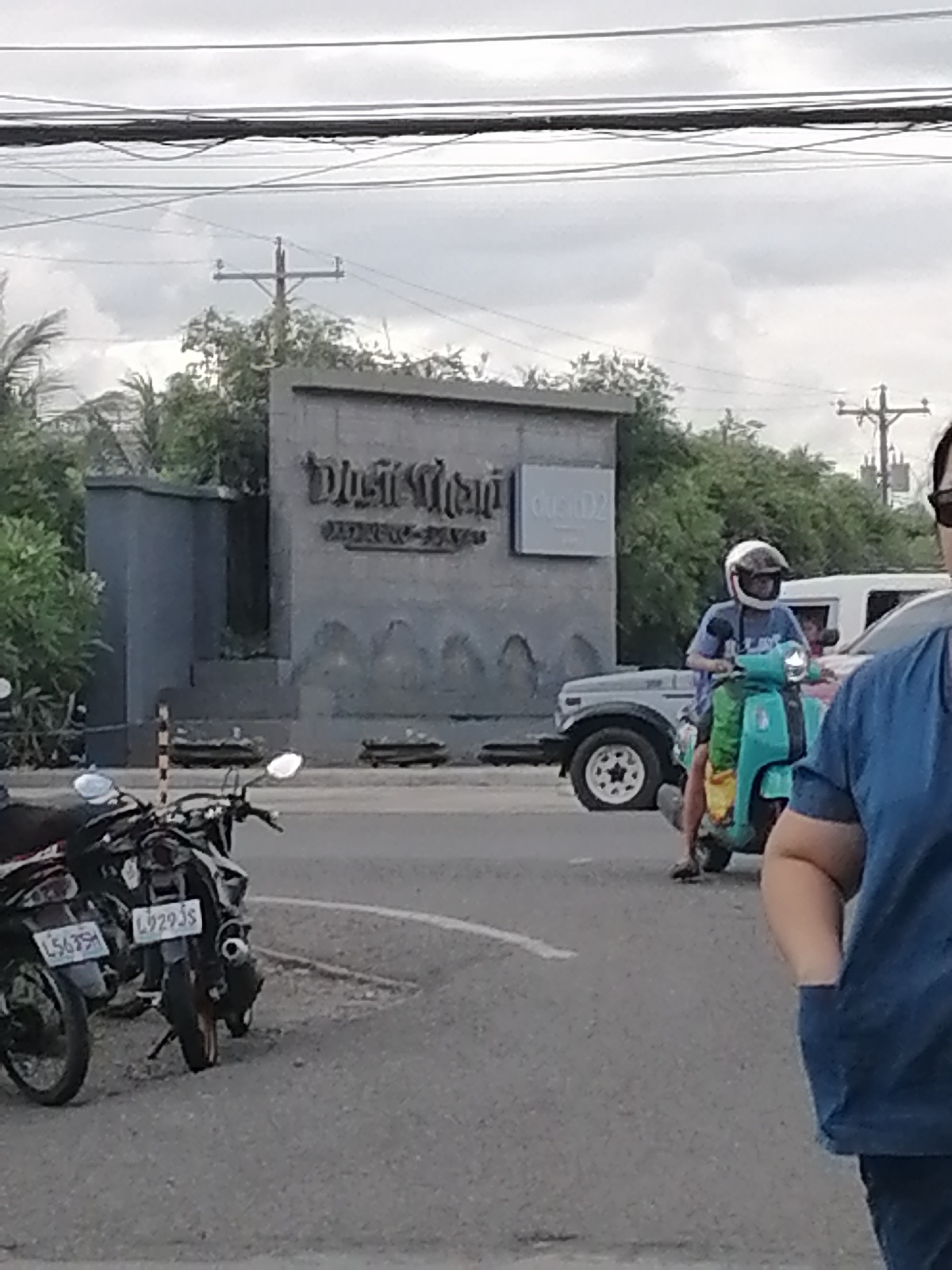 The image depicts an outdoor scene in what is identified as Davao City, Philippines. In the foreground, two motorcycles are parked on the side of a road. One motorcycle, to the left, displays a license plate that is partially visible but appears to read "L5639." The motorcycle to its right has a license plate that reads "L929JS." Behind the parked motorcycles, a person is riding a turquoise scooter, wearing a white helmet and a blue t-shirt. The scooter has a yellow and green accessory attached to the front. Further in the background, a light-colored SUV is parked, and a building with a sign that appears to say "DUST & DIAL" is visible. The sky is overcast with clouds, and power lines are strung across the upper portion of the image. The environment suggests a street or thoroughfare in a built-up area.
