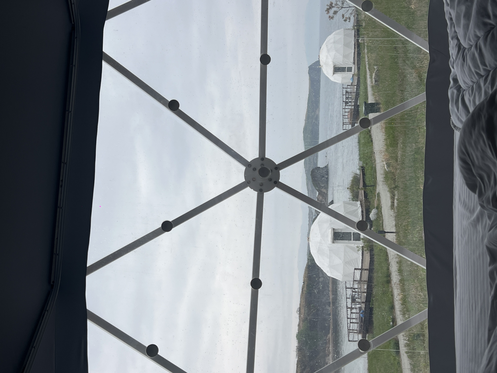 The image shows a view through a dome-shaped structure with a network of white metal bars and circular connectors. The view looks out onto a coastal landscape, where there are two similar dome structures with seating areas, a body of water, a land area with cliffs or hills, and sky obscured by clouds or fog. The location appears to be Zarubino, Russia, based on the presence of distinctive dome-shaped structures. In the foreground, to the left and right of the frame, are the edges of the interior of the dome. The scene suggests it is a tourist or recreational area near the coast.