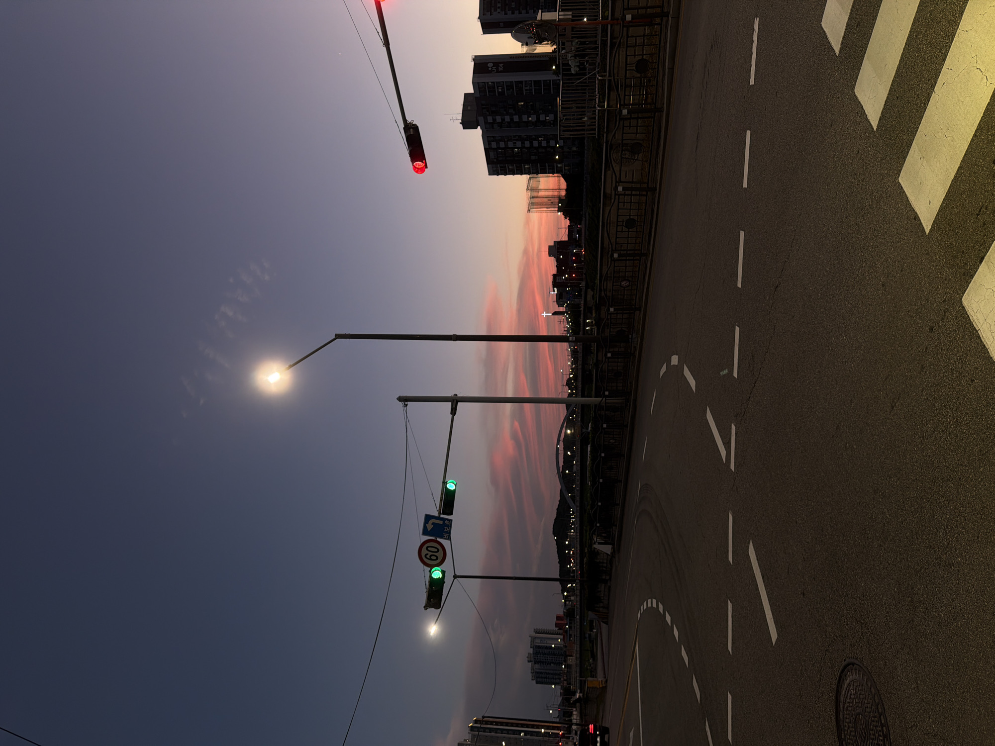 The image depicts a street scene at dusk in Dongducheon-si, South Korea. The sky displays a gradient of deep blue to a soft pink and orange, indicating the transition from day to night. A street lamp illuminates the foreground road, which has white lane markings and a pedestrian crossing. Traffic lights are visible, with one showing a red light and another showing green lights. In the background, several multi-story buildings line the cityscape, and a bridge with an arch spans a body of water. A sign with "60" is also present on one of the traffic light poles. The overall impression is one of urban evening ambiance.