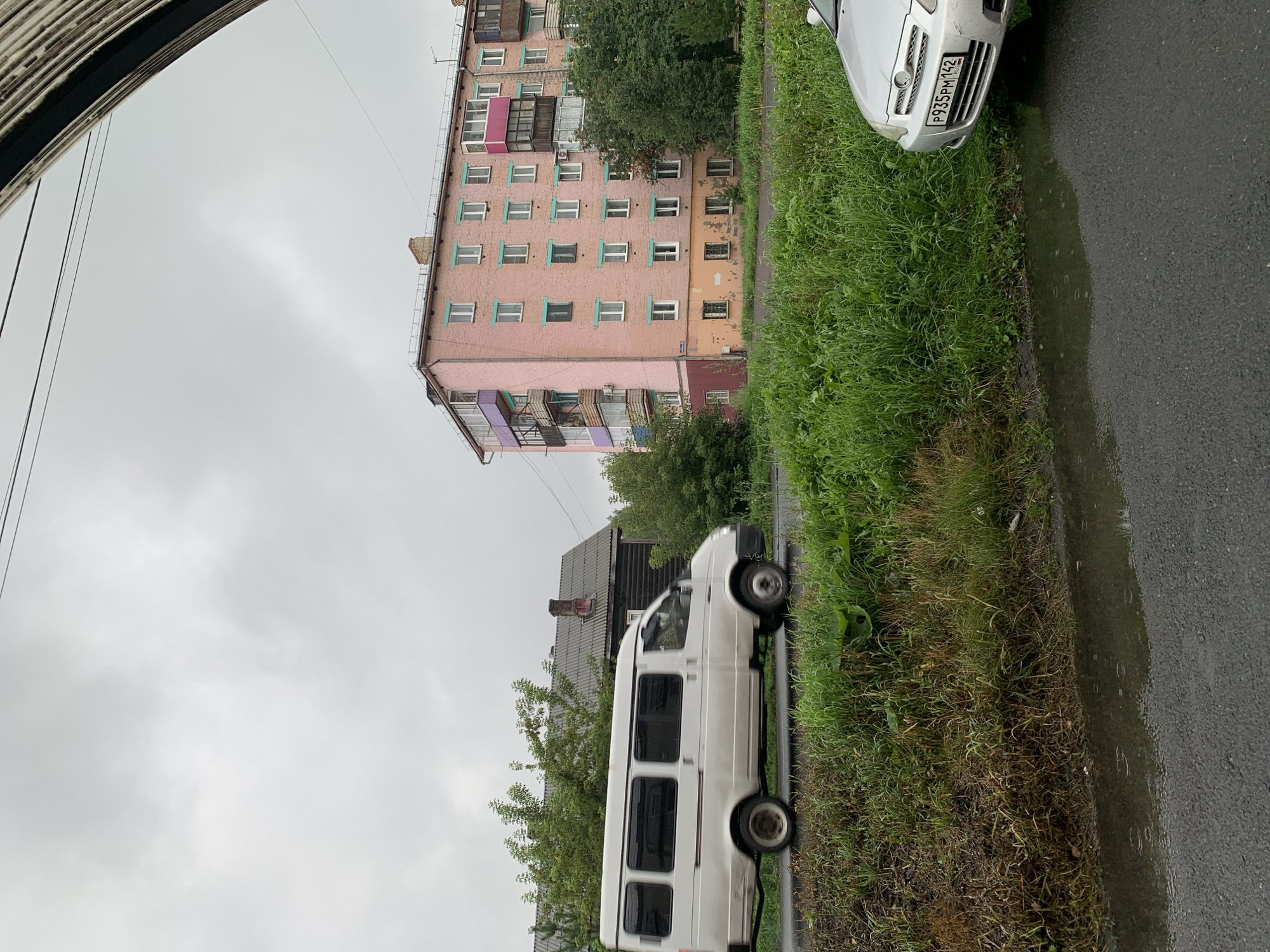 A light pink, five-story apartment building is visible on the right side of the frame in Prokopyevsk, Russia. The building features numerous windows with light blue frames and several balconies, some enclosed with glass and others open. To the left of the apartment building, a dark wooden, two-story house with a grey, gabled roof and a brick chimney stands. In front of these buildings, a white passenger van is captured in motion, appearing to be driving on a wet asphalt road. The van has tinted windows and is positioned to the left of the frame. The foreground is dominated by a roadside embankment densely covered with green grass and some dry, brown vegetation. A puddle of water is present on the asphalt road in the lower part of the image. A silver car is partially visible on the right side of the frame, parked on the road. The sky is overcast and grey, suggesting a cloudy or rainy day. Electrical wires are visible crossing the top of the frame.