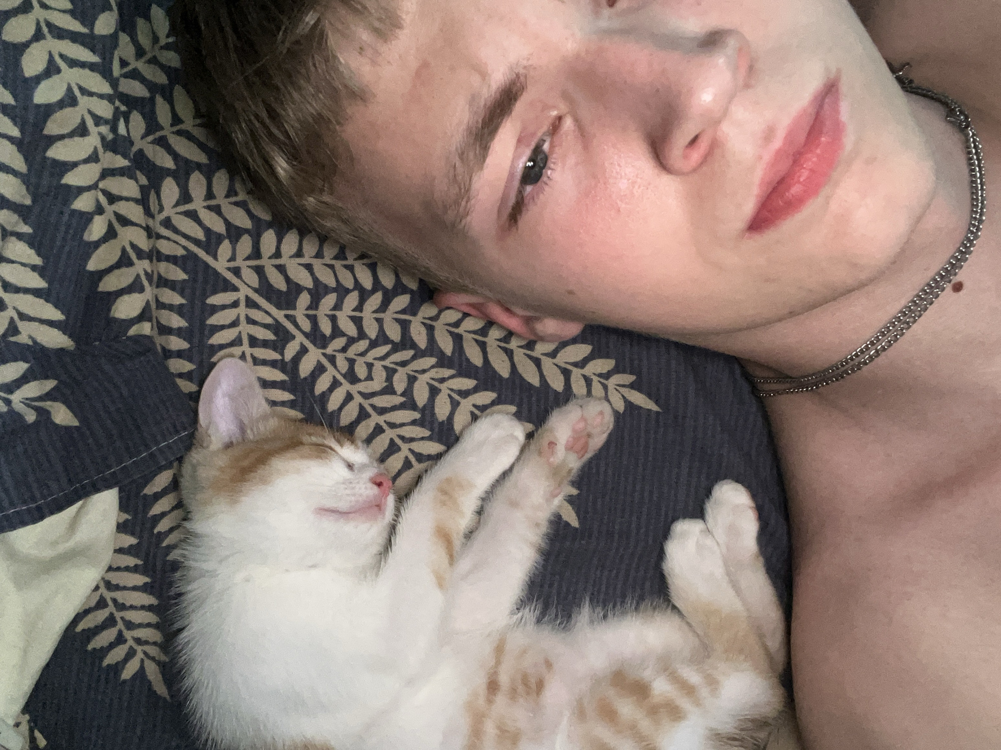 The image shows a close-up, overhead view of a person lying down next to a sleeping kitten. The person's head is tilted back, with their face partially in frame, looking up. The kitten, mostly white with ginger tabby markings, is curled up and sleeping on its back, with its paws extended. Both subjects are lying on a dark blue fabric with a repeating pattern of light beige leaves. The person is wearing a thin, silver chain necklace. The lighting suggests it is daytime, and the overall mood is peaceful and intimate. The scene appears to be indoors, possibly on a bed or sofa. No visible text is present in the image.