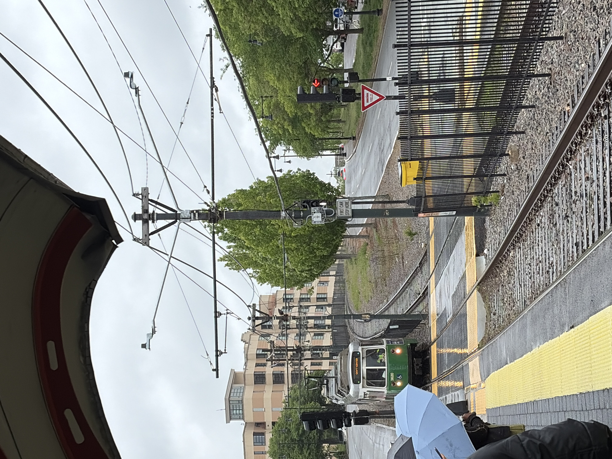 The image depicts a rainy day scene at a platform of the MBTA Green Line, likely the C branch, in Boston, Massachusetts. A green and silver trolley is pulling into a station, with its destination sign reading "GOVT CENTER." Overhead wires supply power to the train. A light blue umbrella is visible, suggesting people are waiting on the platform. A street runs parallel to the train tracks, equipped with traffic lights and yield signs, and lined with trees and adjacent buildings. The overall atmosphere is overcast and wet.