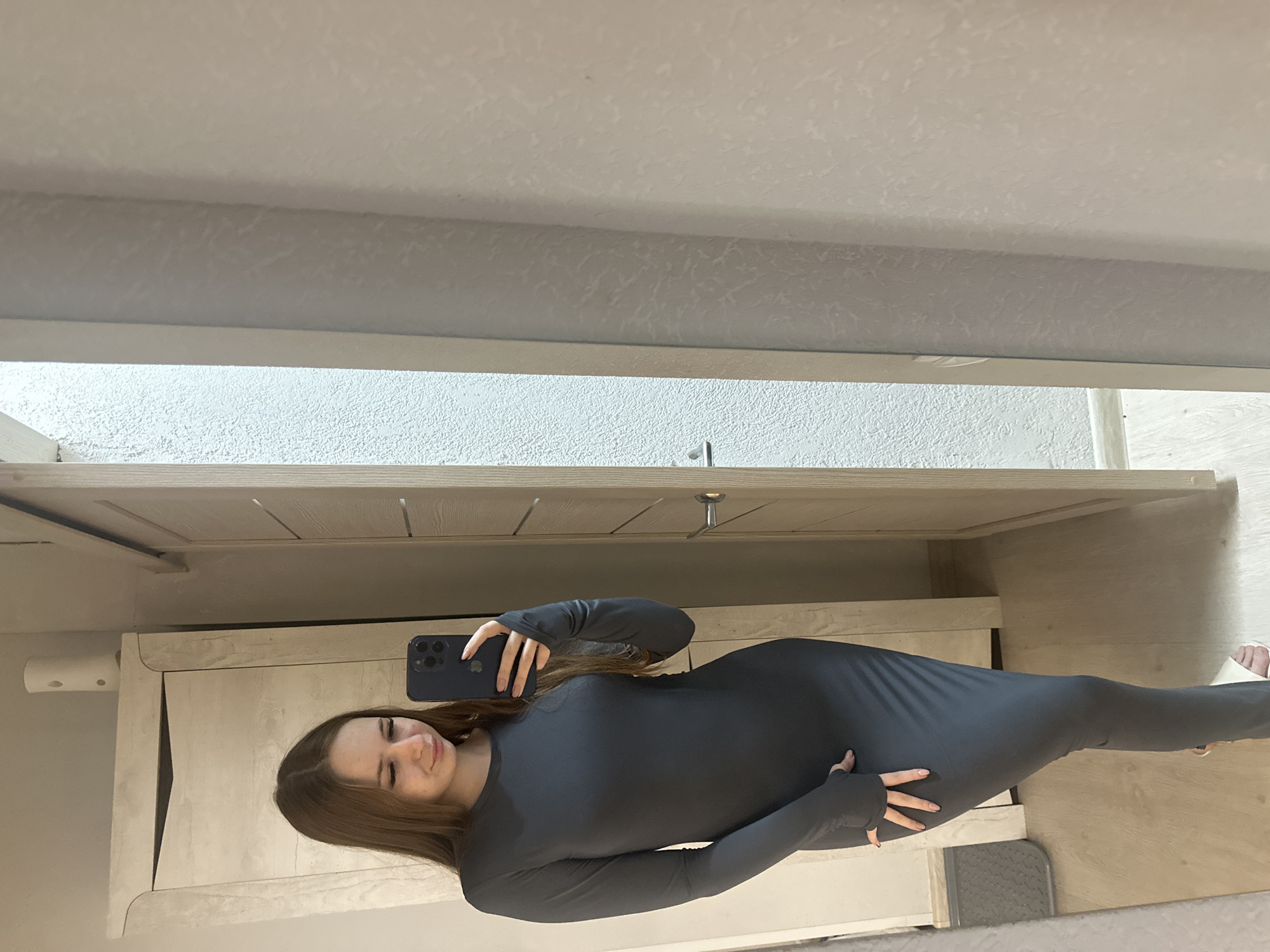 A woman is taking a selfie in a mirror. She is wearing a long-sleeved, form-fitting grey dress. The background shows a room with a white-painted wall and furniture. The location appears to be inside a residence.