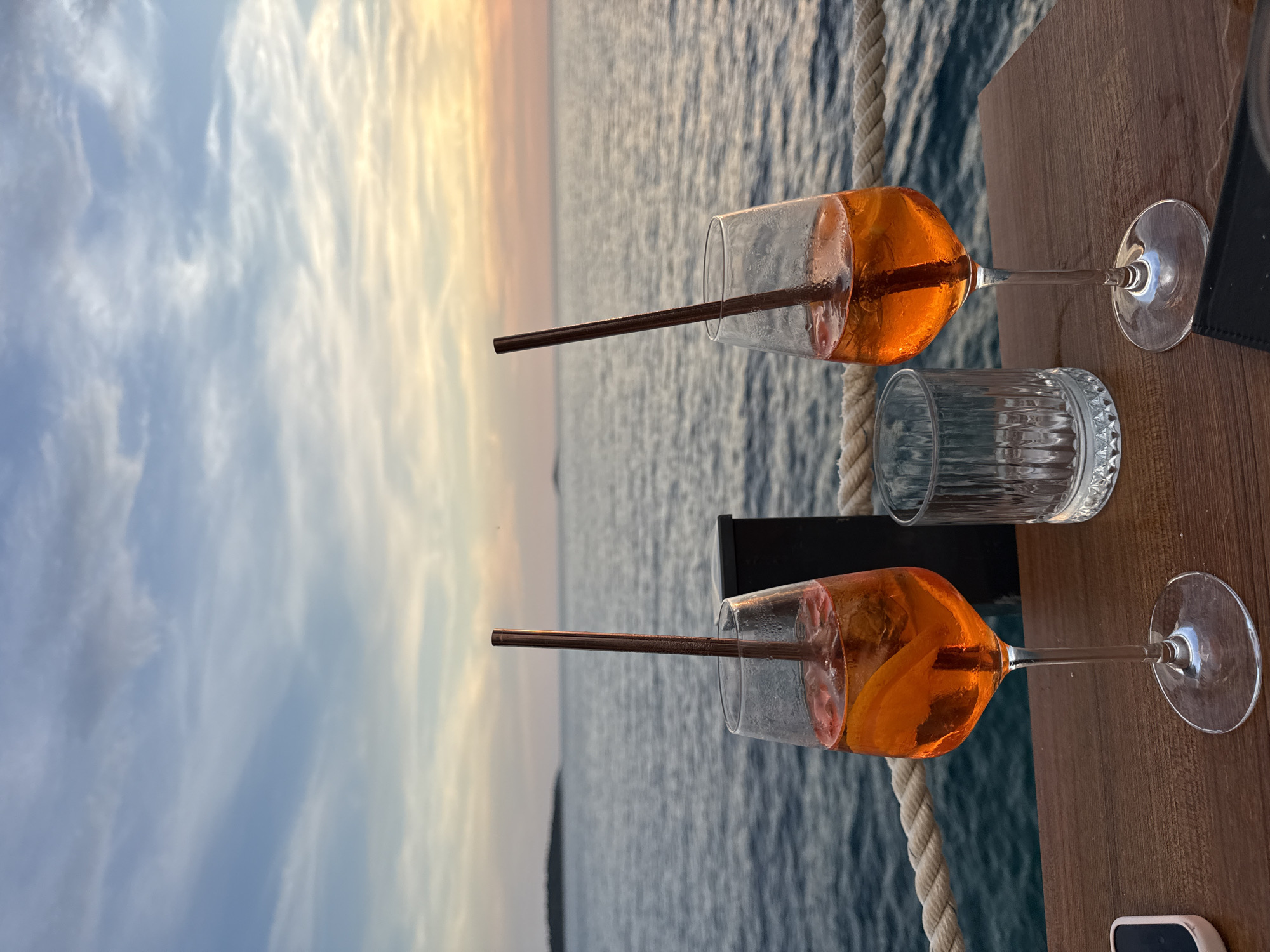Two glasses of orange aperitif drinks with straws sit on a wooden table overlooking the sea at sunset in Primošten, Croatia. The sky is a blend of blue, pink, and orange hues, with wispy clouds. The sea is calm, with gentle waves. In the foreground, a thick rope runs horizontally along the edge of the table. A clear glass with a ribbed texture is positioned between the two drinks. A dark object, possibly a wallet or phone case, rests on the table in the lower right corner.