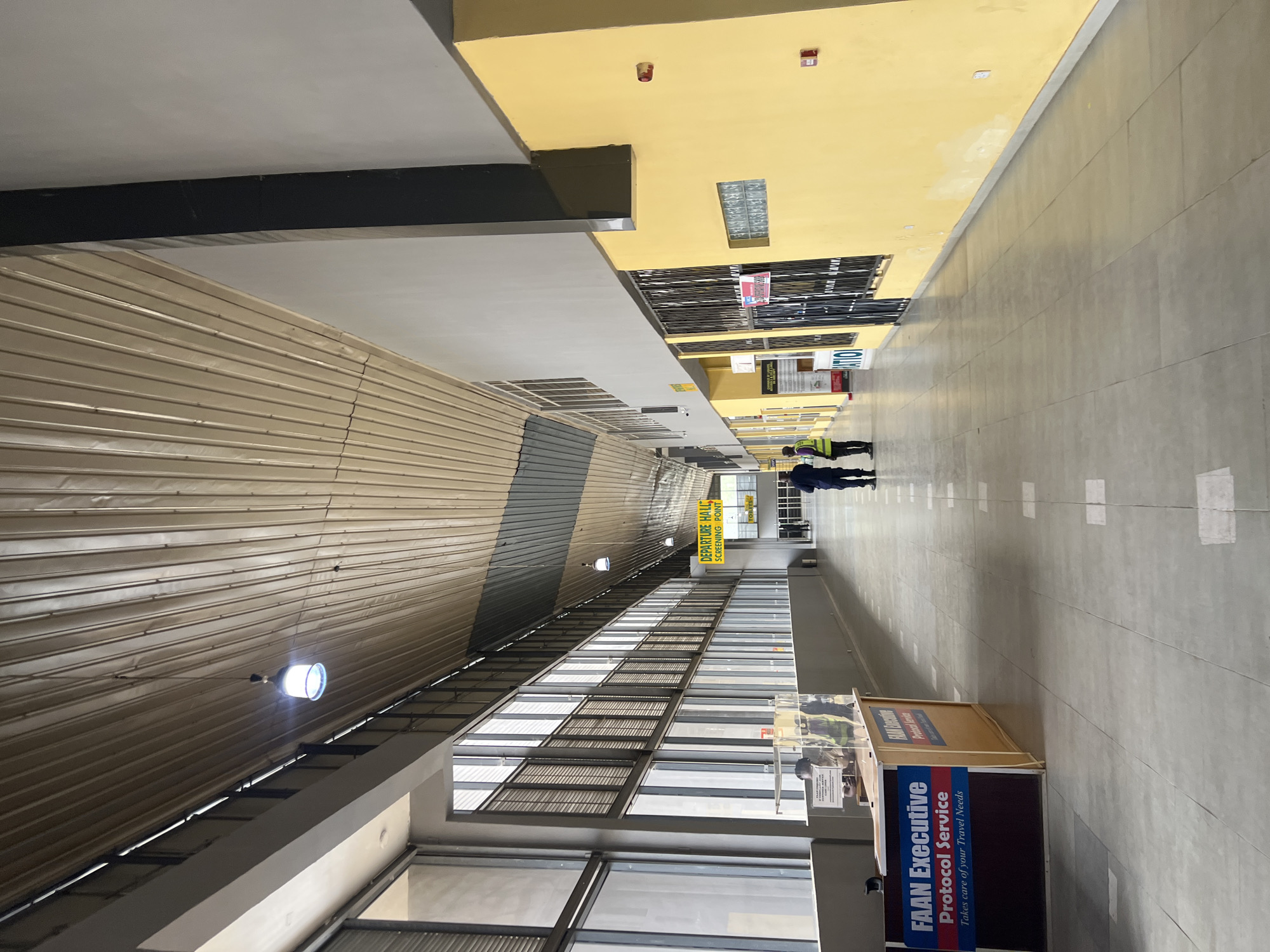 The image depicts an interior space, possibly an airport terminal, located in Kwi, Nigeria. Two individuals wearing what appears to be security vests are positioned further down the hallway. On the right side of the hallway, there is a booth labeled "FAAN Executive Protocol Service." A sign above indicates "DEPARTURE HALL SCREENING POINT." The architecture is marked by a high ceiling, metal siding on one side, and a window-lined wall with multiple panes on the other. The floor is light-colored and appears to be made of tile or a similar material.
