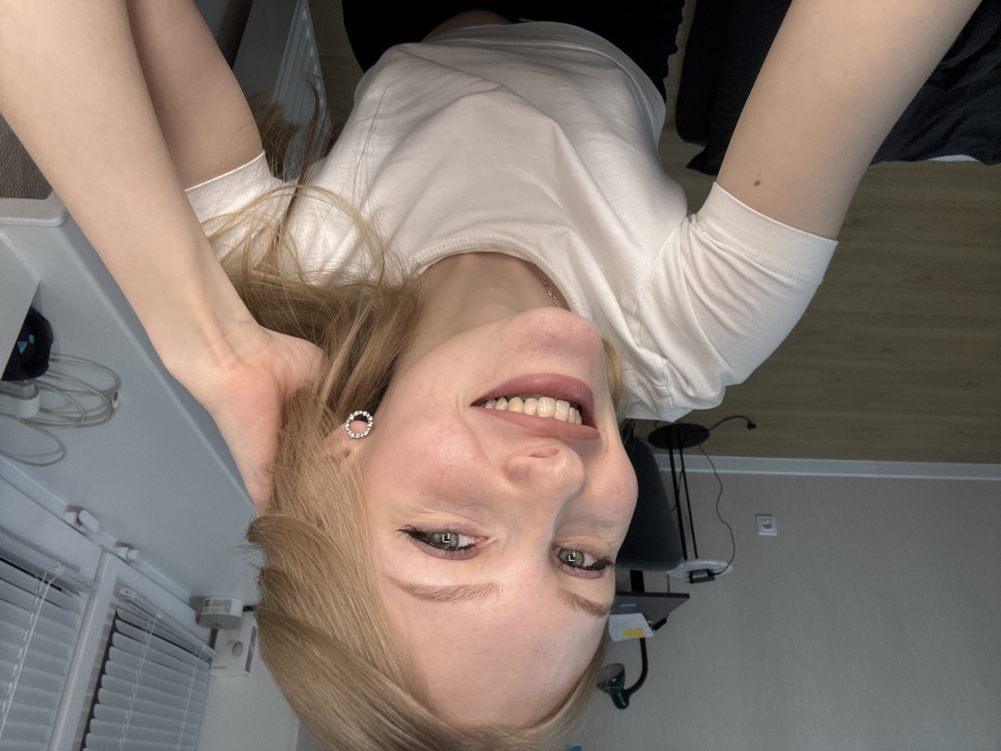 An overhead selfie captures a young woman with blonde hair and light-colored eyes, smiling broadly while looking up towards the camera. She wears a plain white short-sleeved t-shirt and a silver chain necklace. A small, circular, sparkly earring is visible in her left ear. Her expression is joyful and relaxed.
The indoor setting suggests a room, possibly a home office or bedroom, in Perm, Russia. To the left side of the frame (her right side), a white window with horizontal blinds is visible, along with a white windowsill holding some electrical wires and a dark object. A white rectangular device is mounted on the wall above the blinds.
To the right side of the frame (her left side), a dark piece of furniture, possibly an office chair or desk, is partially seen, along with a dark-colored lamp with a flexible arm. The background includes a light gray wall with a white electrical outlet. The lighting appears to be artificial, making the exact time of day difficult to determine. No discernible text is visible in the image.