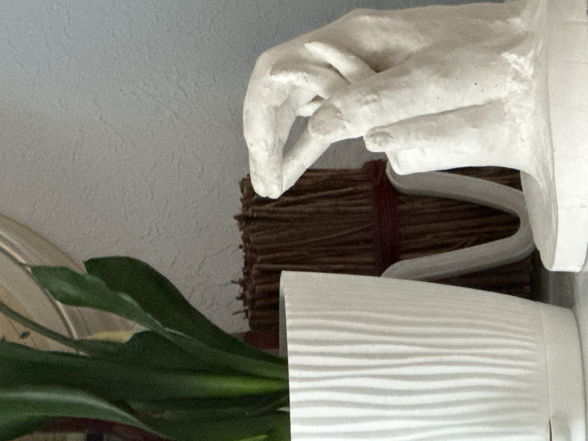 The image shows a close-up of a white textured plant pot, a bundle of brown dried reeds tied with red string, and a white plaster sculpture of hands. The plant pot has vertical ridges and contains green leaves. The reeds are arranged in a bundle. The sculpture depicts two hands, one with fingers extended and the other with the thumb and index finger touching. The background is a textured light blue wall. The scene is indoors, and there is no specific indication of its location within the city of Nevinnomyssk, Russia. The lighting suggests it is daytime.