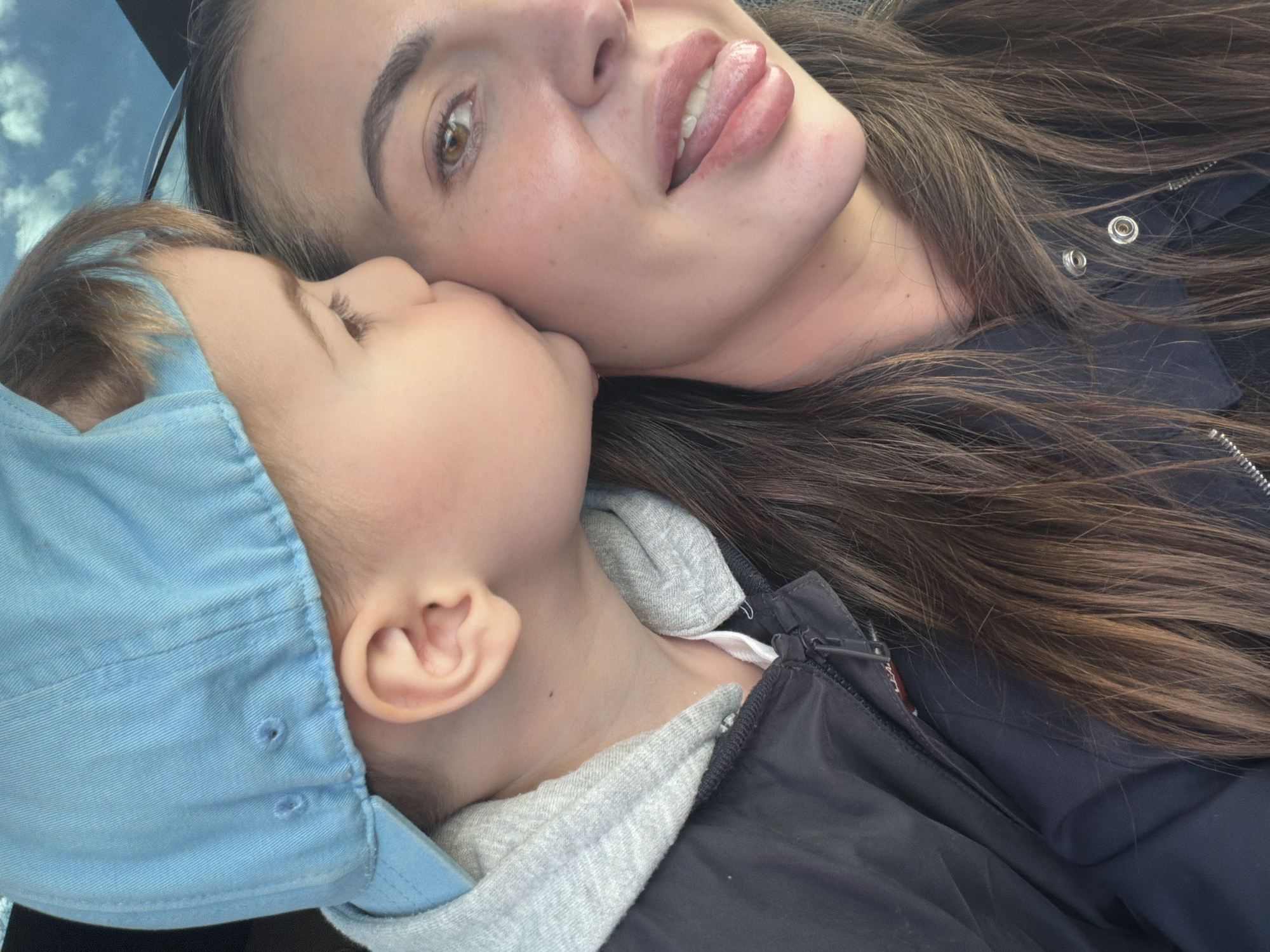 The media file is a close-up, overhead portrait capturing an intimate moment between a woman and a young child, likely a mother and son. They appear to be lying down, possibly taking a selfie, with a bright, cloudy blue sky partially visible in the upper left background, indicating a daytime setting in Krasnoyarsk, Russia.
The woman, positioned on the right side of the frame, has long, dark brown hair that spreads around her head. She has fair skin, light brown eyes, and full lips with her tongue playfully extended. She is wearing a dark-colored jacket, possibly black or dark blue, with metallic snap buttons visible on the lapel. A pair of sunglasses rests partially on top of her head.
The child, on the left, is snuggled close to the woman's face and neck, facing away from the camera. The child's light brown hair is visible beneath a light blue baseball cap, which features subtle stitching and small air holes. The child wears a gray hooded sweatshirt layered under a dark, zippered jacket, similar in style to the woman's. The child's ear and a portion of their neck are visible.
The image conveys a warm and affectionate atmosphere, highlighting the close bond between the two individuals. A small red tag with white text, likely a brand label such as "LEVI'S", is visible on the zipper pull of the woman's jacket.