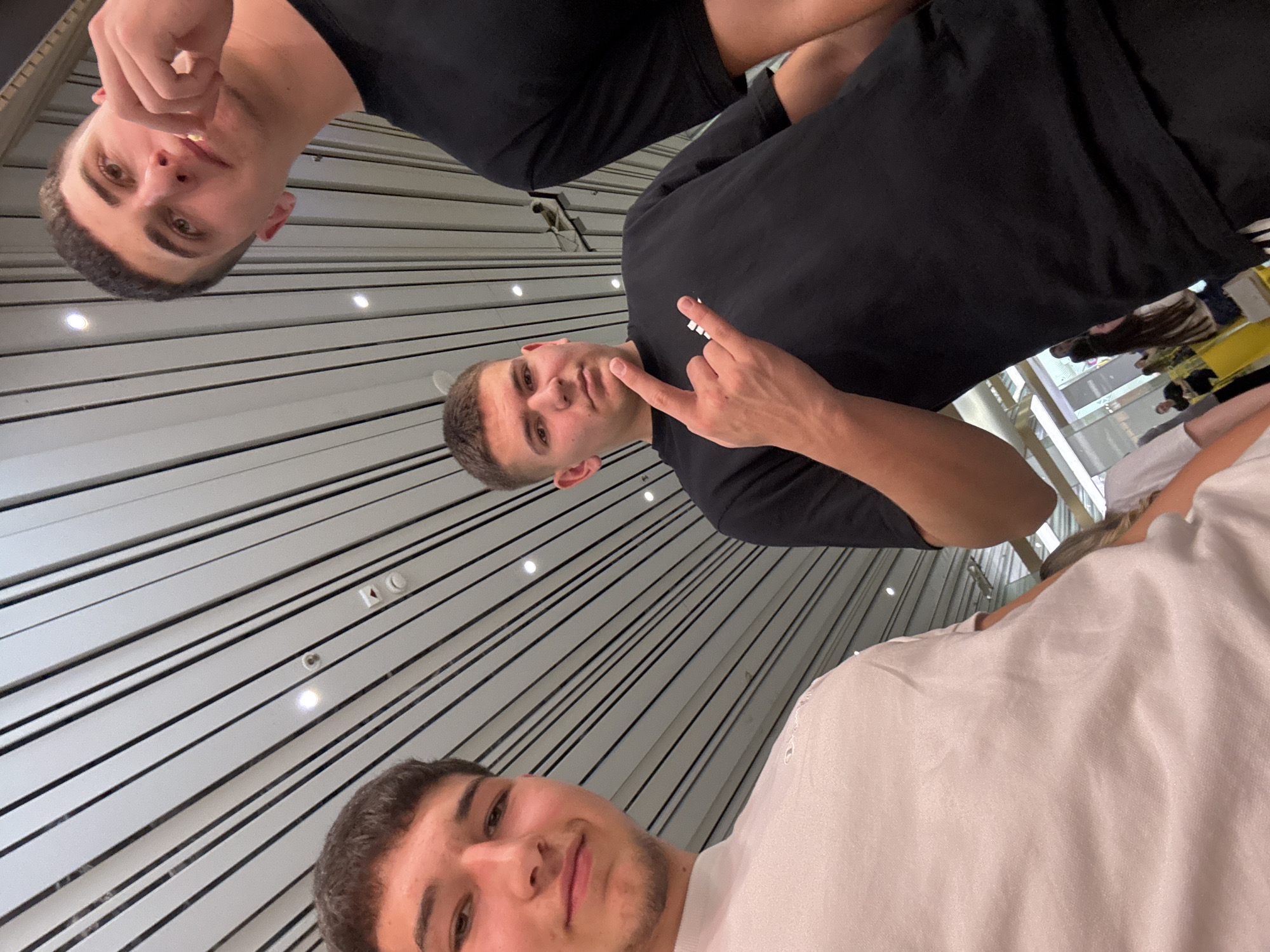 Three young men are taking a selfie. The man at the top of the frame has short, dark hair and is wearing a black t-shirt. He is looking down at the camera and making a gesture with his hand. The man in the middle is also wearing a black t-shirt and has short, dark hair. He is pointing at the camera. The man at the bottom of the frame is wearing a white t-shirt. The background appears to be a ceiling with parallel white slats and recessed lighting. There are other people and objects visible behind them, seemingly in a shopping center. It is difficult to confirm the location is specifically in Plovdiv, Bulgaria, without additional information.