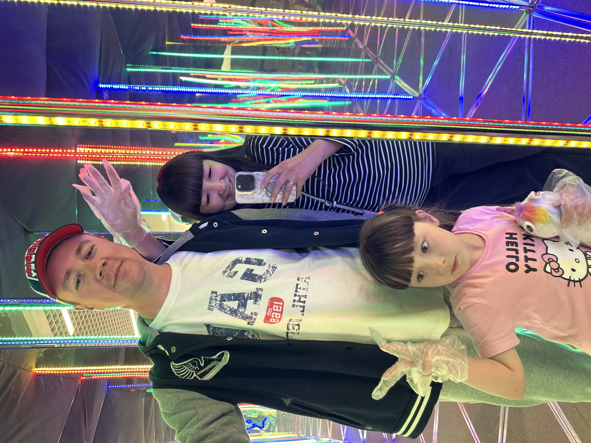 In Izhevsk, Russia, a family of three is taking a reflective selfie in what appears to be an amusement park or funfair attraction, possibly an "Infinity Mirror" room, characterized by numerous vertical LED light strips in red, yellow, blue, and green. A Caucasian man, wearing a black and grey bomber jacket over a white t-shirt with blue lettering and a red and black baseball cap, stands on the left, holding up his right hand, covered by a clear plastic glove, in a peace sign gesture. To his right, an East Asian woman is taking the selfie with her phone, wearing a black and white striped shirt and a gold ring on her left ring finger. In the foreground, a young Caucasian girl with dark hair and bangs, wearing a pink t-shirt with "HELLO YTTIK" printed in white, is held by an adult, her right hand also covered by a clear plastic glove, making a peace sign. She is holding a stuffed toy. The environment is dark with the dominant lighting coming from the numerous reflections of the LED strips creating a tunnel effect.