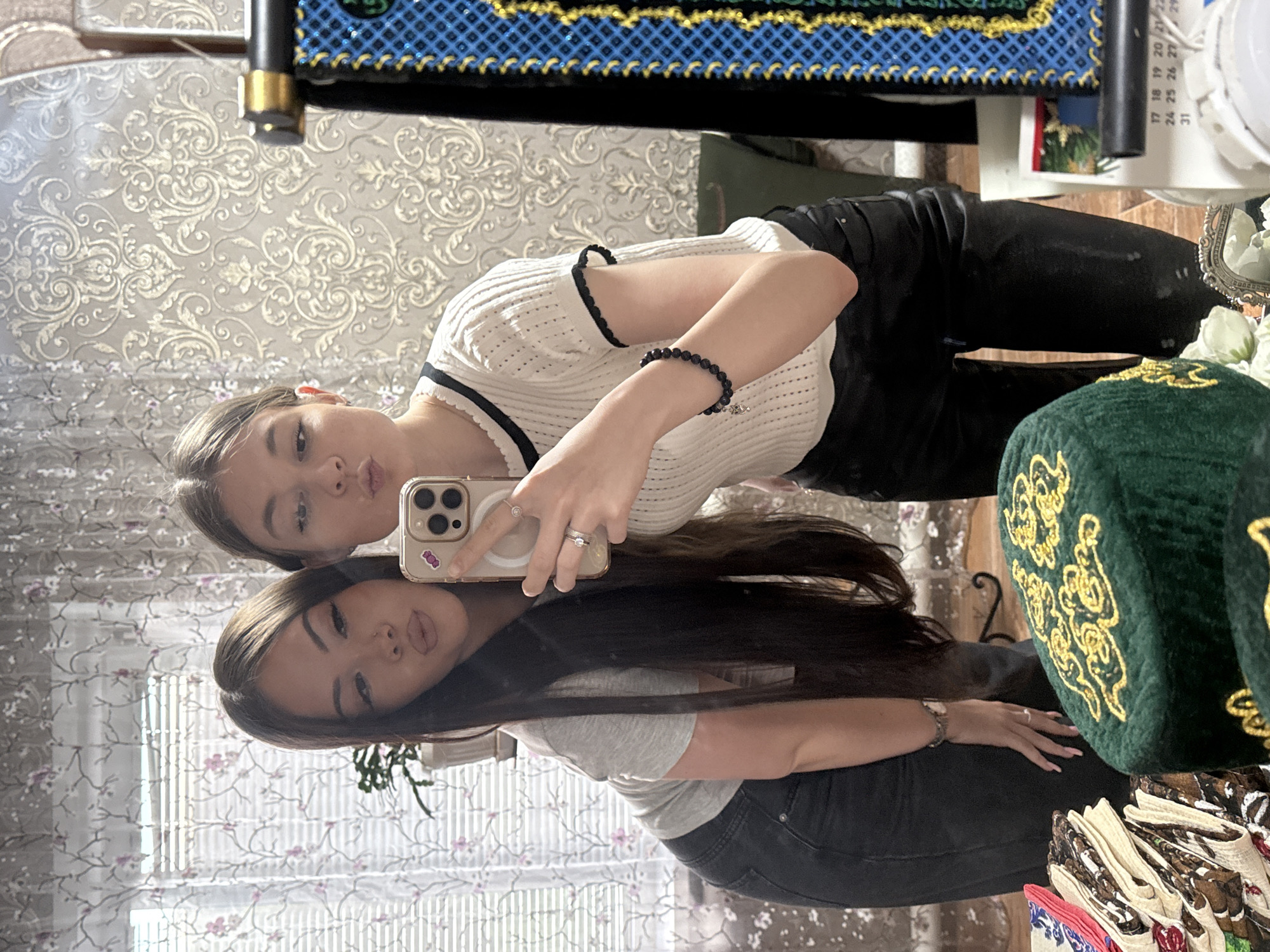 A mirror selfie of two young women, reflecting the room. One is wearing a white, short-sleeved shirt with black trim, black pants, and a black beaded bracelet. The other is wearing a grey shirt and dark jeans. Each is making a duck face. A phone is in the upper woman's hand. Other items visible in the reflection include a green embroidered hat, patterned fabric, and a calendar. The wallpaper is light with a floral pattern. The location is Коноваловка, Russia.