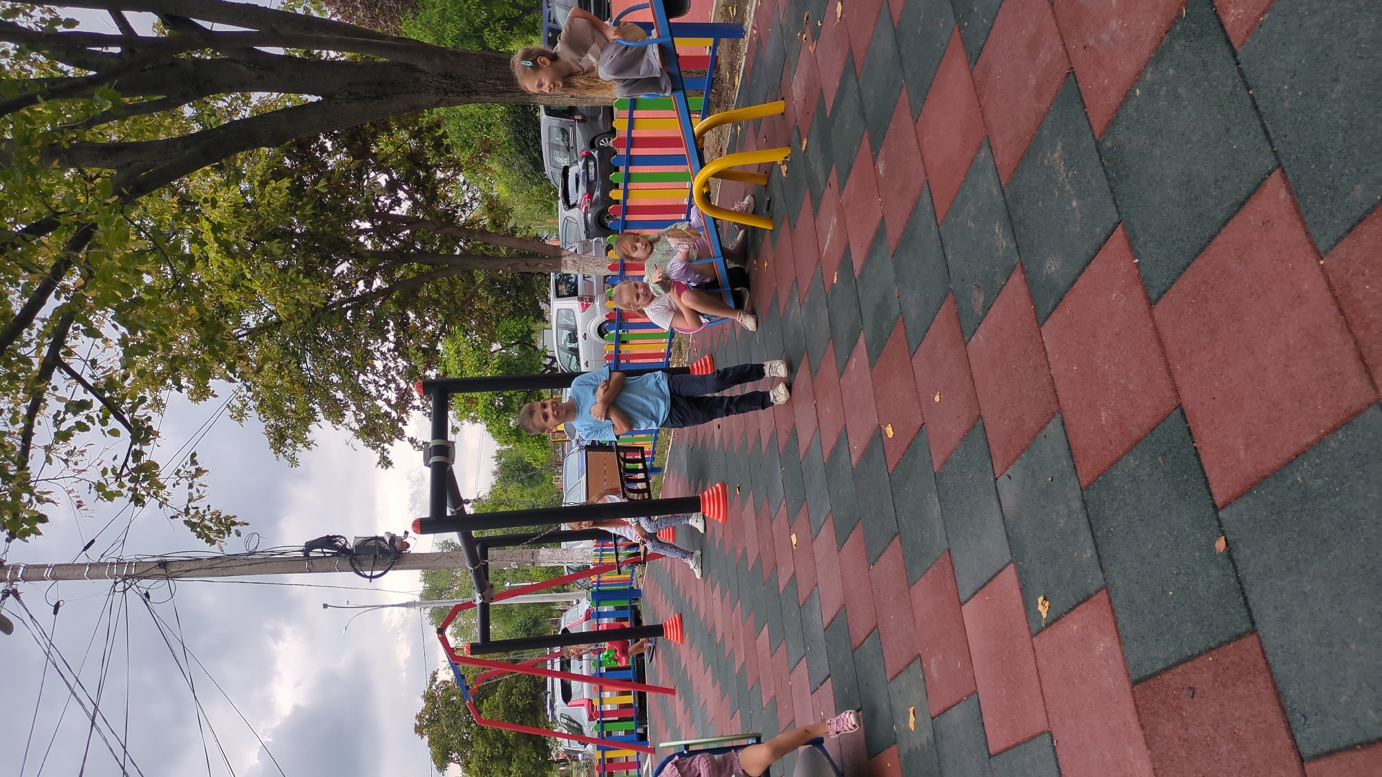 The image depicts a playground with a rubberized surface composed of red and dark grey tiles. In the foreground and mid-ground, several children are present. A boy in a blue t-shirt and dark pants stands with his arms crossed, looking towards the viewer. To his right, two young girls are seated on a yellow seesaw. Further back, a girl in a white shirt sits on a swing set. Behind the playground, a colorful fence separates the area from a street with parked cars. Trees with green and yellowing leaves are visible in the background, and a utility pole with numerous wires stands prominently on the left. The sky is overcast. Based on the presence of specific brand markings on vehicles visible in the background, such as a Dacia Logan, and the general architectural style of the surrounding buildings (partially visible), this playground is likely located in Chișinău, Moldova.