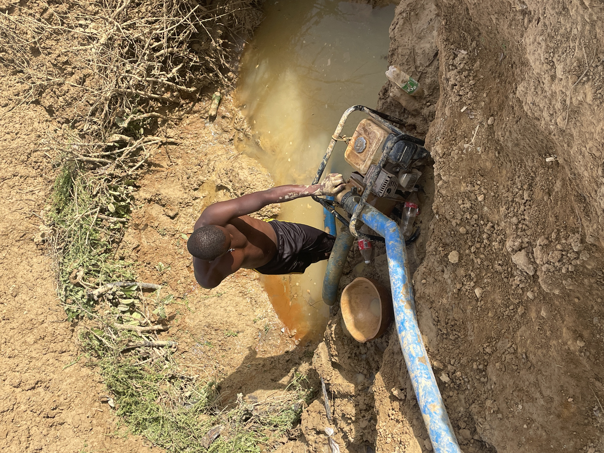 In an outdoor environment near Gana Daji, Nigeria, an individual with dark skin, wearing black shorts and no shirt, stands beside a muddy hole filled with murky water. The person appears to be operating a gasoline-powered water pump, indicated by the hoses and the visible engine. The pump sits close to the edge of the hole. A wooden bowl rests on the ground next to the blue pump hose. The immediate surroundings include soil, sparse vegetation, twigs, and discarded plastic bottles. The landscape appears dry and dusty.