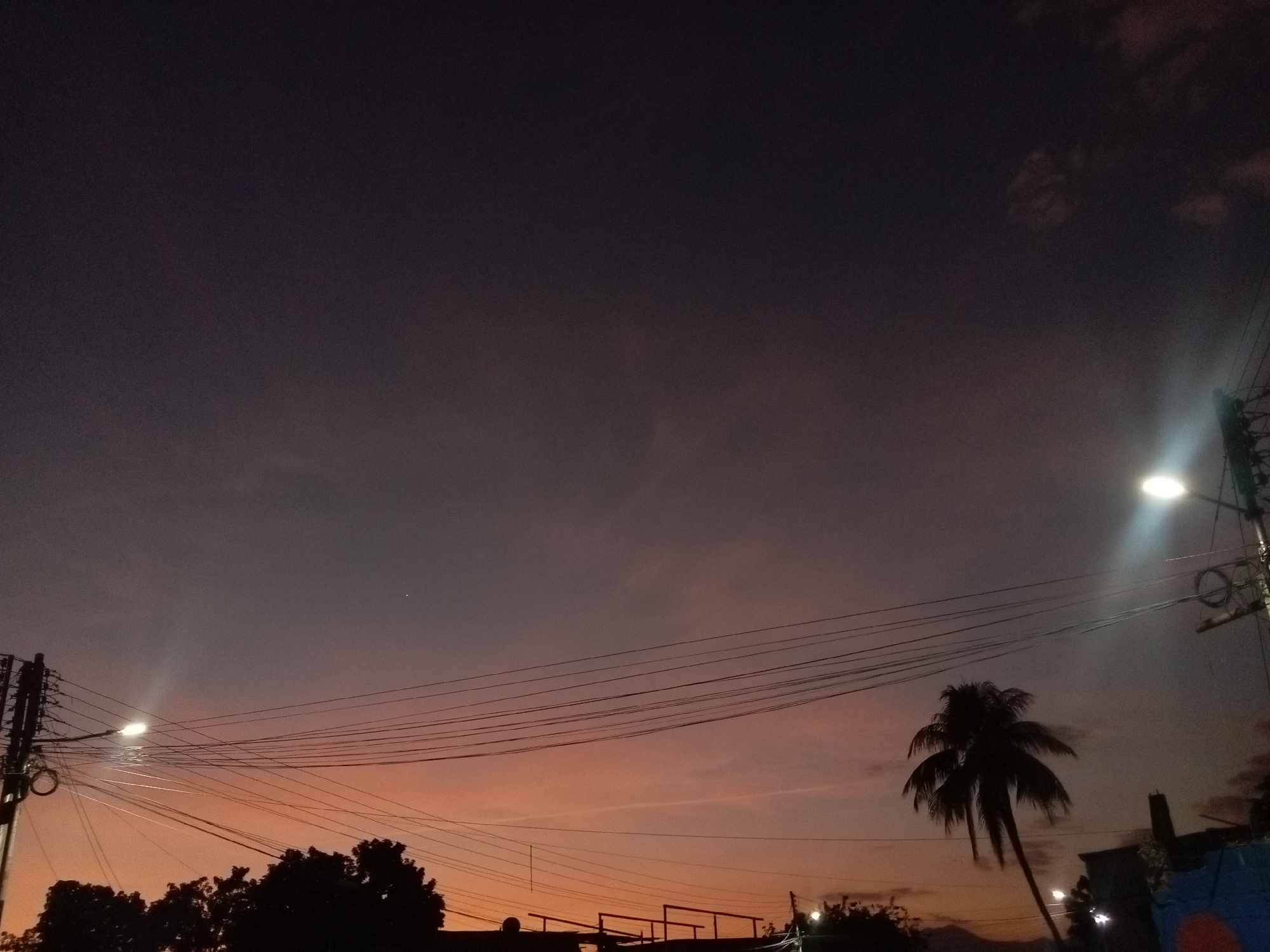 The image depicts a dusk or twilight scene in Cagua, Venezuela. The sky transitions from a dark purple-gray above to orange-pink near the horizon. Silhouettes of trees and a palm tree are visible against the lower sky. Power lines crisscross the view. Streetlights illuminate the scene, one on the left and another on the right. The lower portion of a building or structure is visible. There are no people visible.