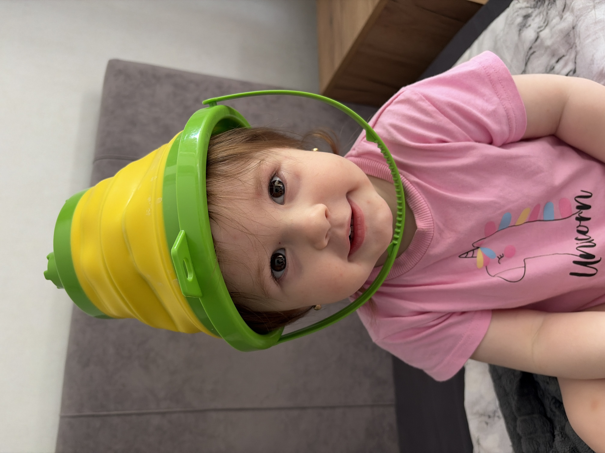 A young child, likely a toddler, is captured in a close-up, upward-facing shot, lying down indoors. The child has light brown hair and dark eyes, looking directly at the camera with a wide, happy smile, showing several small teeth. A bright yellow and green collapsible plastic bucket is placed upside down on the child's head, with the green rim framing their face like a hat. A green handle from the bucket extends forward over the child's forehead.
The child is wearing a short-sleeved pink t-shirt adorned with a colorful unicorn graphic and the word "Unicorn" written in black cursive text. The visible portion of the child's arm is bare.
The background suggests a domestic setting. A grey upholstered surface, possibly a sofa or headboard, is visible behind the child's head, contrasting with a light-colored wall. On the right, a patterned grey and white duvet or blanket is visible, alongside a wooden surface. The lighting indicates it is daytime. The image conveys a sense of innocent playfulness and joy within a home in Chaikovsky, Russia.