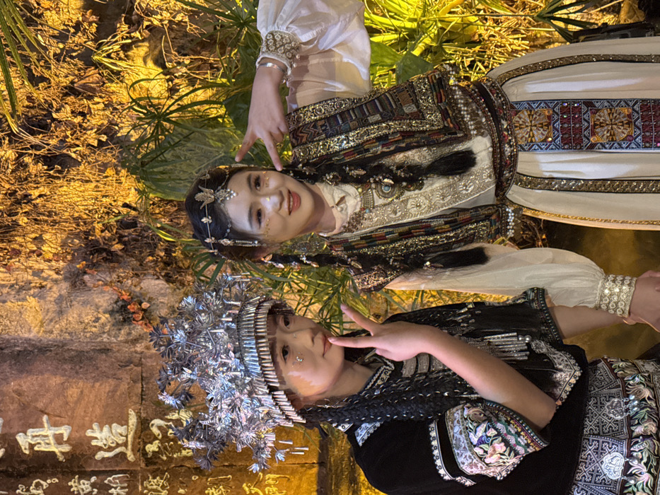 This image captures two young women posing for a photograph at night in what appears to be an outdoor setting, possibly a park or cultural site in Yuzhong District, China. Both women are adorned in elaborate traditional attire, characteristic of certain ethnic minority groups.
The woman on the left wears a striking, multi-tiered silver headpiece that resembles traditional Miao (Hmong) headwear, along with a dark, richly embroidered outfit featuring silver ornaments and intricate patterns on the sleeves and bodice. She poses with a slight smile and holds up her hand in a peace sign. Gold-colored embellishments are visible on her face.
The woman on the right is dressed in a cream-colored blouse with puffed sleeves, covered by a vest heavily embroidered with vibrant gold and multicolored threads. Her head is adorned with a more delicate, gold-toned headpiece featuring floral or butterfly motifs, and her hair is styled in long braids. She has a wide smile and makes a "rock on" hand gesture. She also has subtle facial embellishments, possibly glitter or small jewels.
The background is softly lit by warm artificial lights, illuminating lush green foliage, creating a glow around the subjects. Behind the woman on the left, a stone wall with carved Chinese characters is visible, though the text is partially obscured and not clearly legible. The overall scene suggests a festive cultural event, performance, or a themed photo shoot, emphasizing the beauty and richness of their traditional garments. Both women appear cheerful and engaged.