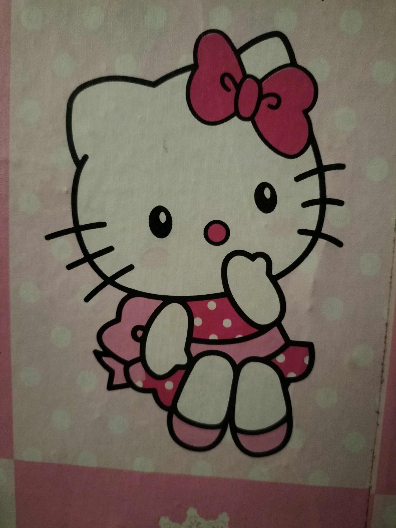 A close-up, eye-level, full-body shot shows a cartoon illustration of Hello Kitty. The character is depicted in a frontal view, seated and looking directly at the viewer. Hello Kitty has a white face with a pink bow on her head, black eyes, a yellow nose, and black whiskers. She is wearing a pink polka-dot dress with a ruffled hem and a pink bow at the neckline. Her hands are raised to her mouth, and her feet are visible below the dress, wearing pink shoes. The background is a light pink fabric with white polka dots. The image appears to be a print on a textile material, possibly a blanket or a tapestry. The lighting is even, suggesting an indoor setting. There are no people present in the image. The location of Sambirejo, Indonesia, is not discernible from the image itself.