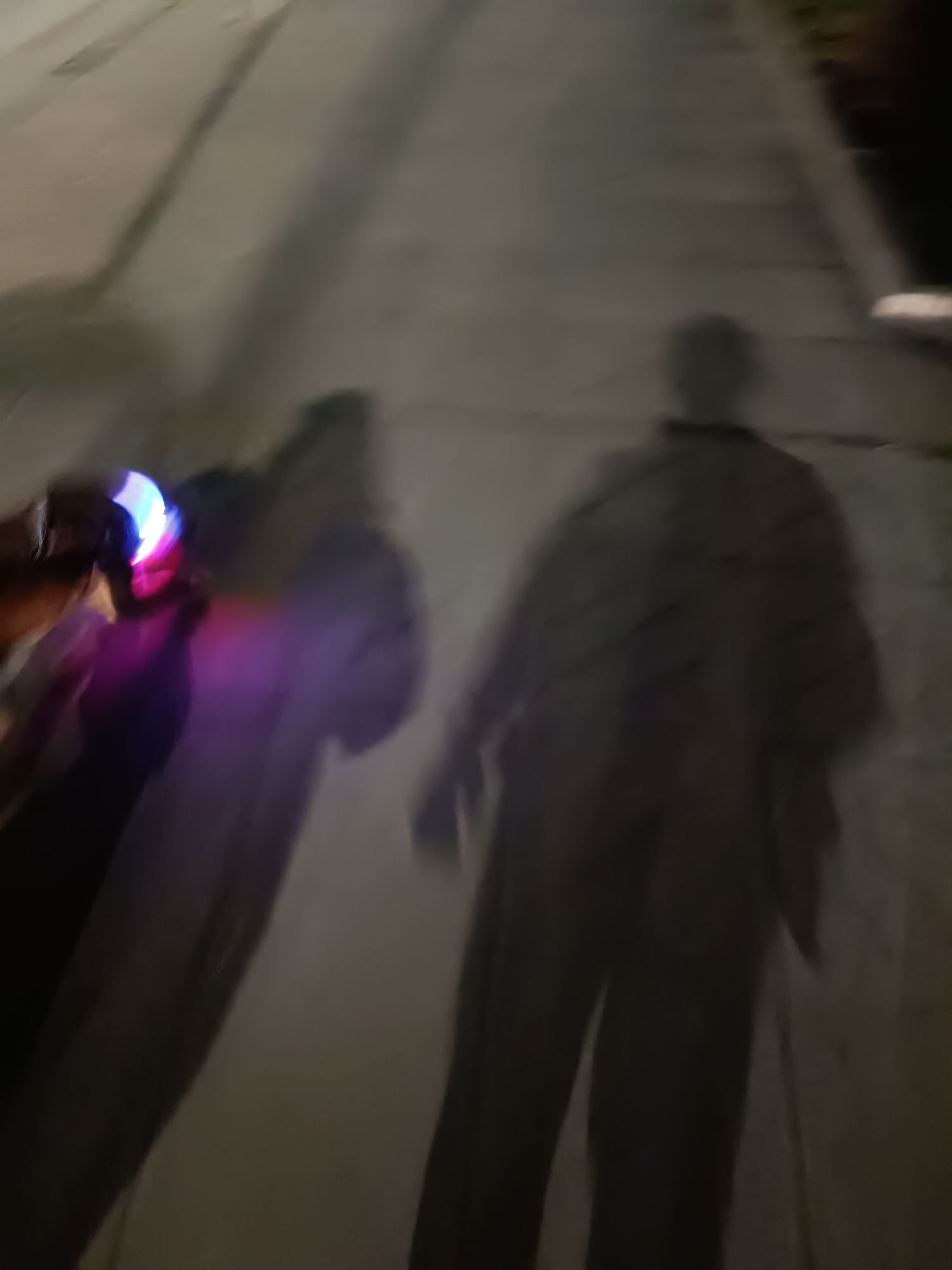 The image appears to be a blurry, low-light photograph taken on a sidewalk or paved area. A person holding an object emitting colorful light (possibly a toy or decoration with blue, pink, and white colors) is visible in the foreground. The shadow of the person is also cast onto the sidewalk, along with another shadow resembling a human figure. The environment is indistinct, but the presence of a sidewalk suggests an urban or suburban area. There is no definitive information to place the scene in Pleven, Bulgaria.