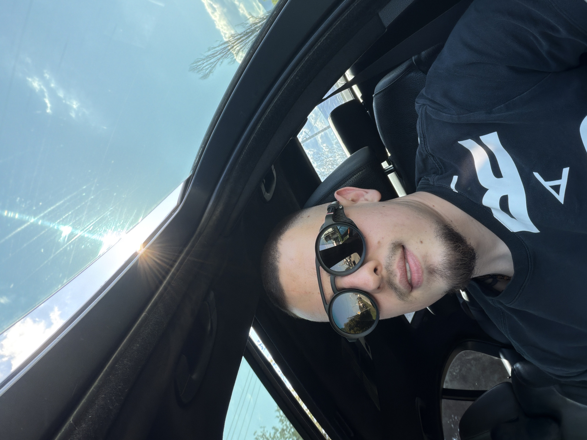 A man with short dark hair, a beard, and sunglasses is inside a vehicle with a sunroof. The sun is shining through the sunroof, casting a glare on the windshield. The interior of the car is black. The man is wearing a black t-shirt with white writing. The location is unknown; Byaga, Bulgaria is not discernible from the image.