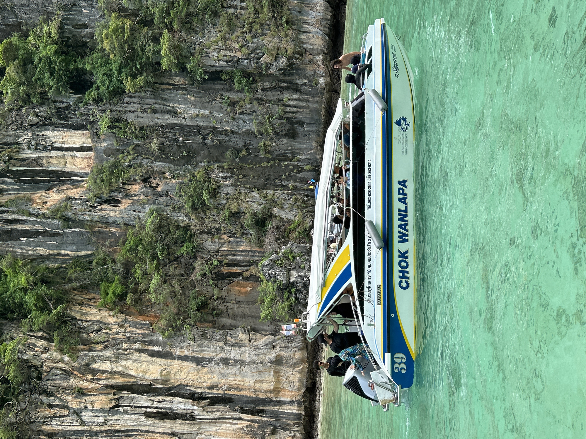 The image shows a speed boat with "CHOK WANLAPA" written on its side, numbered "39", in the turquoise waters off the coast of Ao Nang, Thailand. The boat, which appears to be a passenger ferry, has a white canopy and is adorned with blue and yellow stripes. Several people are visible on board, some standing and others seated under the canopy. In the background, a towering limestone cliff covered in lush green vegetation rises dramatically from the water. The water is clear and calm, with sunlight dappling its surface. The overall scene depicts a typical day of island hopping or sightseeing in the popular tourist destination of Ao Nang.