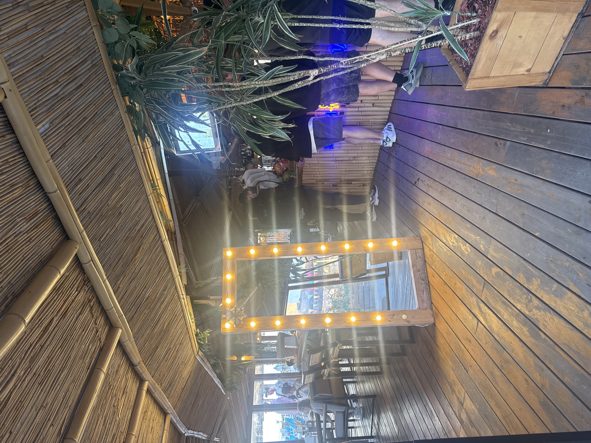 The image shows a partially outdoor setting with wooden plank flooring and a bamboo-covered ceiling. A large, illuminated mirror frame is prominent in the center, reflecting a scene of people seated at tables inside, with a bright, sunlit view visible through windows. Several people are standing or walking around in the outdoor area. A potted plant with long, green and white striped leaves is visible near the top left. There are blue neon lights underneath the wooden planks in the flooring. This picture appears to be taken in a cafe or bar, the specific location is in Sirius, Russia.