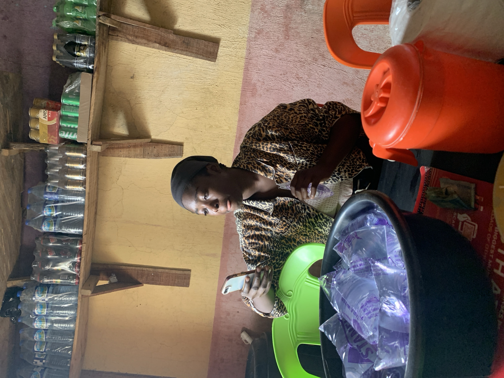 A woman sits on a green chair in what appears to be a small store in Rafin Par, Nigeria. She is wearing a leopard-print shirt and a black head covering. She holds a cell phone in one hand and a sachet of water in the other. A black tub filled with more water sachets sits next to her. Behind her, an orange water container is visible. A rudimentary wooden shelf is on the wall, stocked with bottled and canned drinks. The walls are painted in two tones, yellow and pink.