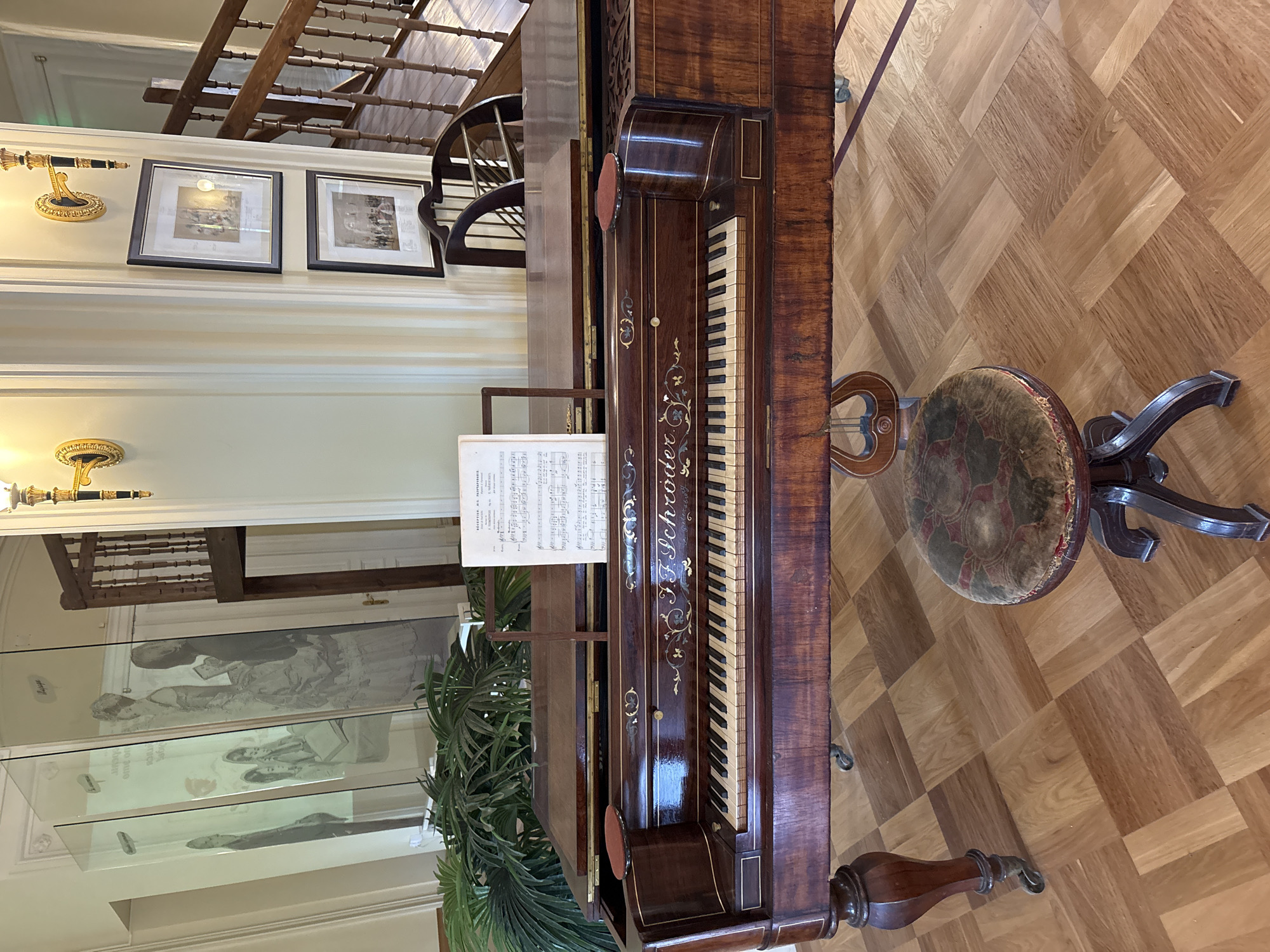 The image shows an interior scene, likely a museum or historical building. In the foreground, a dark wood antique piano is positioned on a parquet floor. Sheet music is displayed on a stand above the keyboard, which features ivory and ebony keys. A vintage piano stool with a patterned velvet seat is in front of the piano. To the left of the piano, there are several tall glass display cases containing historical drawings and text, possibly depicting people in period attire. Behind the piano and display cases, a wooden staircase with a railing ascends to an upper level. On the wall to the right of the staircase, two framed pictures are hung. A potted plant with large green leaves is visible to the left of the piano. The overall setting appears to be well-lit with ambient light and decorative wall sconces. This scene is located in the Gatchina Palace, Gatchina, Russia, specifically in the Music Room.