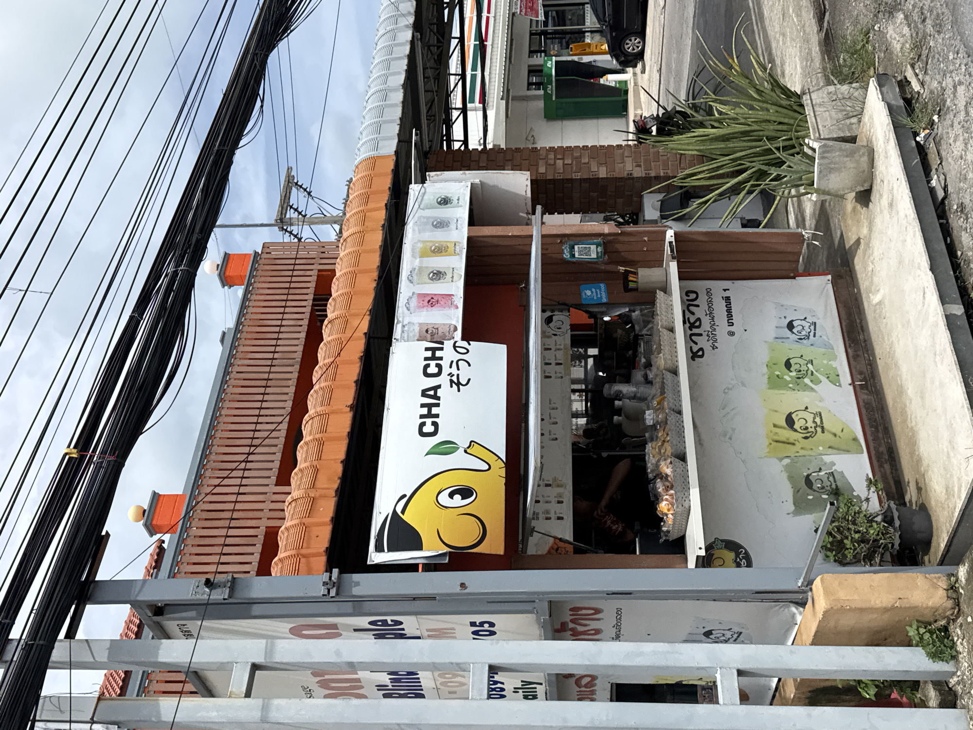 The image shows a small outdoor food stall in Rawai, Thailand, specializing in beverages. The stall features a prominent sign with a yellow elephant mascot and the text "CHA CHA". Beneath this, there are colorful images of drinks. The stall has a counter with various items displayed, including baskets of pastries and containers of utensils. Two potted plants, one a snake plant and the other a smaller succulent, are situated in front of the stall. In the background, power lines are strung overhead. Further in the distance, a 7-Eleven store and a black car are visible. The overall scene depicts a typical street view of a commercial area in Rawai.