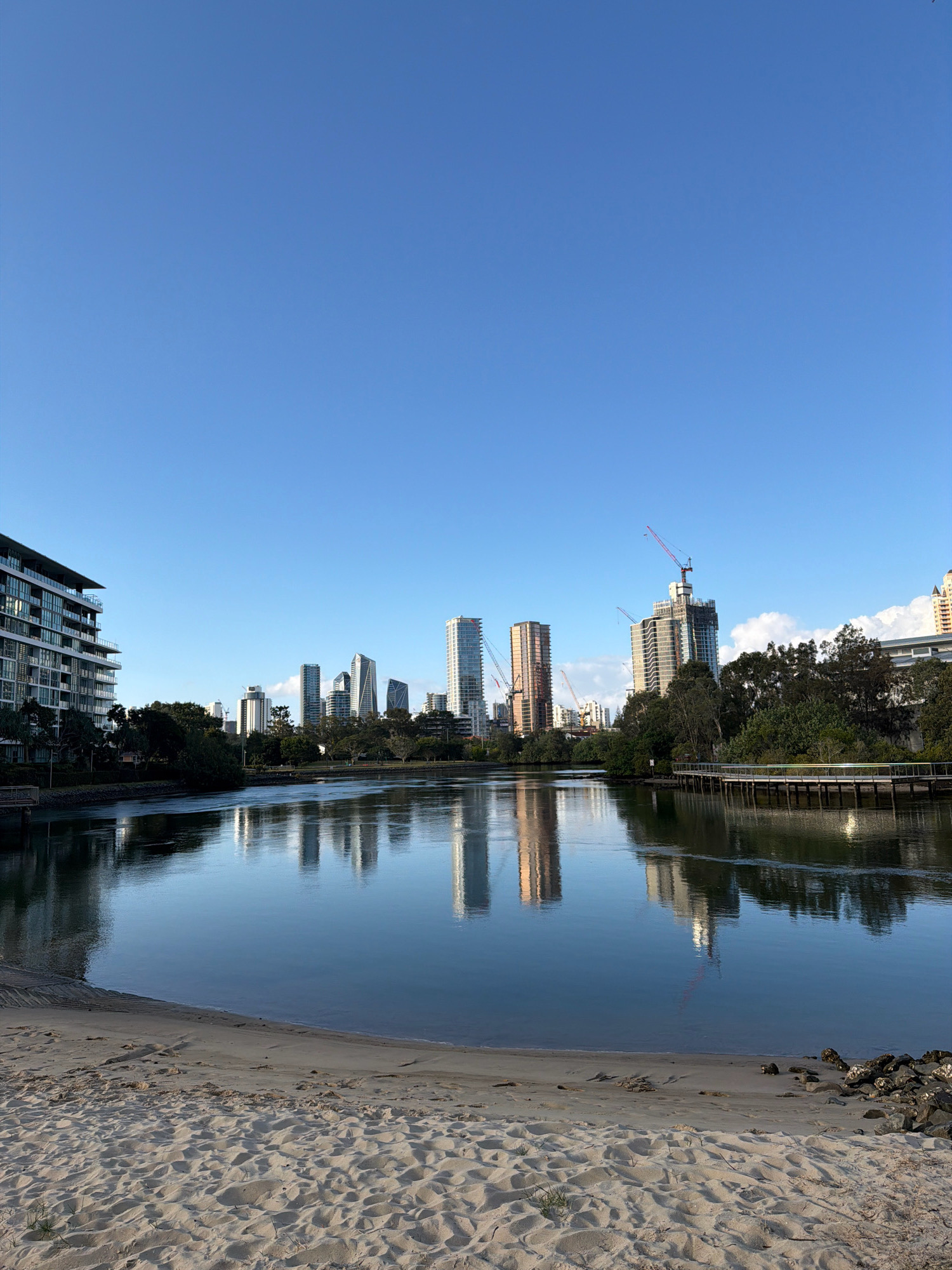 This is a wide-angle, eye-level, outdoor photograph of a tranquil river scene in Broadbeach Waters, Australia, on a clear, sunny day. The foreground features a sandy beach with some scattered rocks and sparse vegetation. The middle ground is dominated by a calm, reflective river, mirroring the surrounding buildings and trees. The background showcases a cityscape with numerous modern high-rise buildings, some of which appear to be under construction, indicated by cranes. Lush green trees line the riverbanks, separating the water from the urban structures. The sky is a clear, bright blue, with no clouds visible. The overall ambiance is peaceful and serene, capturing the confluence of nature and urban development.