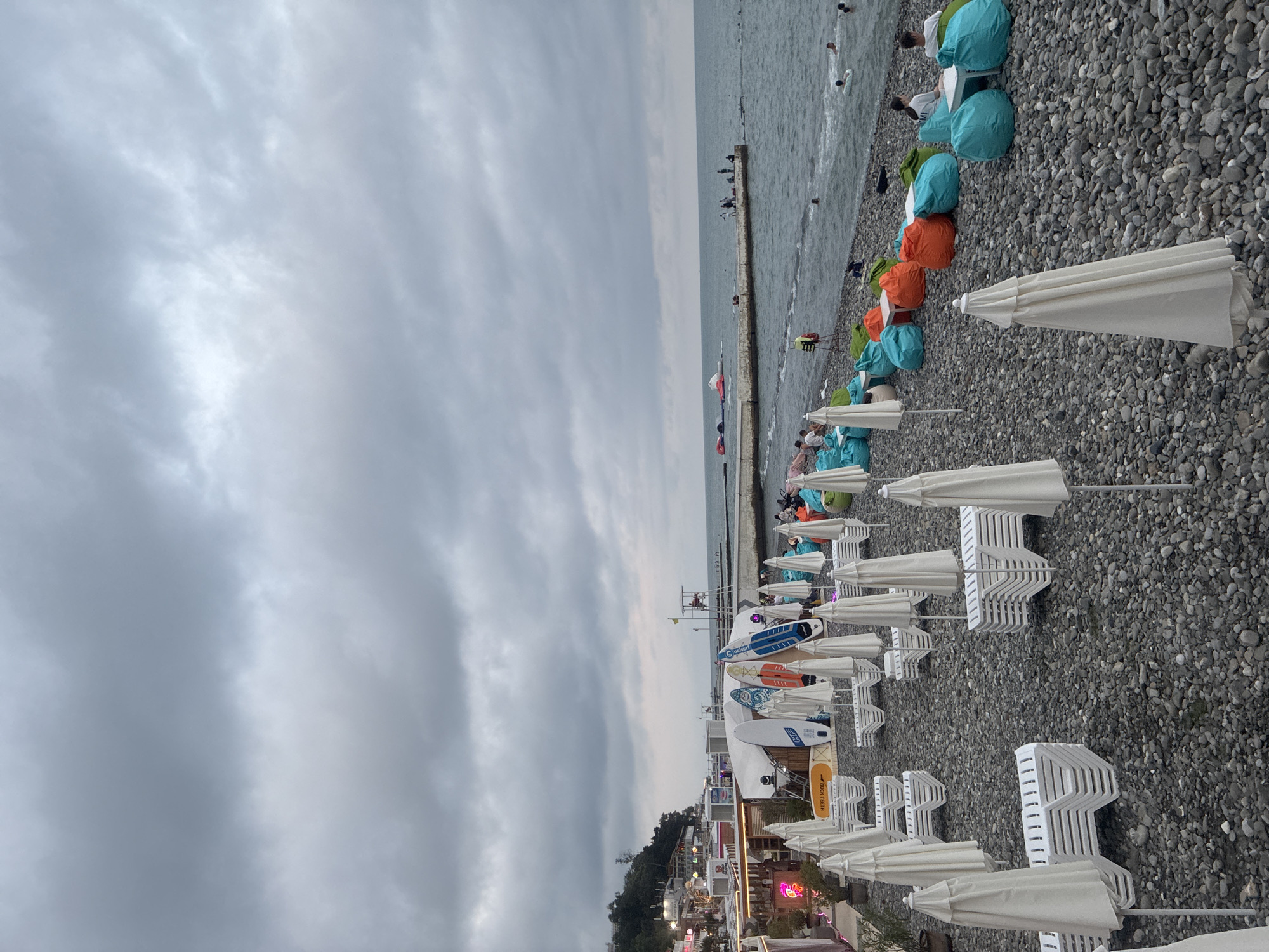This is an exterior, eye-level shot of a pebble beach on a cloudy day, likely in Sochi, Russia, given the context. The scene is sparsely populated, with the focus on rows of white beach chairs and closed white umbrellas neatly stacked and arranged on the grey pebbles. Further down the beach, near the water's edge, are several colorful beanbag chairs in shades of orange, green, and turquoise. A few people are seated on these beanbag chairs, and some are in the water. In the middle ground, a concrete pier extends into the sea, with a few individuals standing on it. Surfing equipment, including paddleboards, is leaning against a white tent structure on the left side of the frame, adjacent to a small, brightly lit building. In the distance, more buildings and a lush green hillside are visible under the overcast sky. The sea is calm with gentle waves.