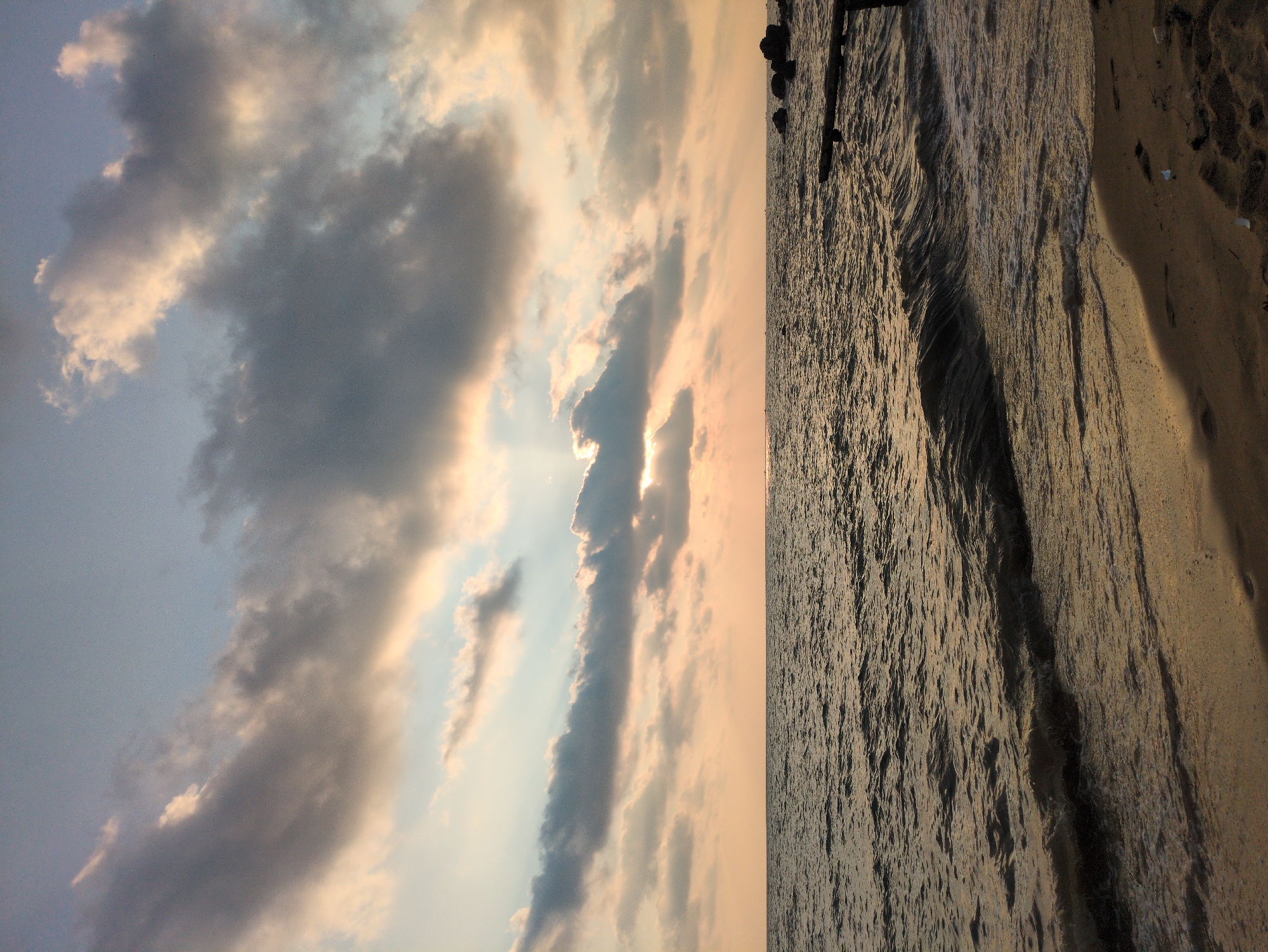 The image shows a horizontal view of a seascape with a cloudy sky during sunset. The sky is filled with clouds, some of which are dark gray, while others are light gray or white. The sun is visible through a gap in the clouds, creating a bright light effect. The ocean is dark and has ripples. On the right side of the image, there is a strip of sandy beach. There are no people visible in the image. The scene appears to be at the city of Phú Quốc, Vietnam.