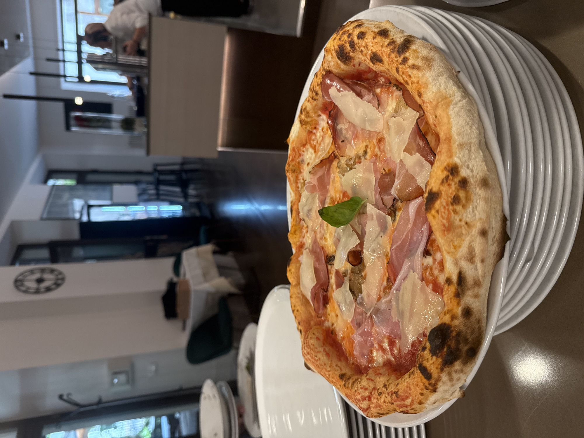 A close-up, eye-level shot features a freshly baked Neapolitan pizza resting on a stack of white plates. The pizza boasts a golden-brown, blistered crust and is topped with tomato sauce, mozzarella cheese, thinly sliced prosciutto, and shaved Parmesan. A single basil leaf garnishes the center. In the background, the interior of a restaurant is visible. A person in a white chef's uniform is partially visible behind a counter to the right. Tables with white tablecloths and chairs are set in the middle ground, with windows and doors leading to an outdoor area or another part of the establishment. A large clock adorns the wall. The overall setting suggests a dining establishment in Trento, Italy.