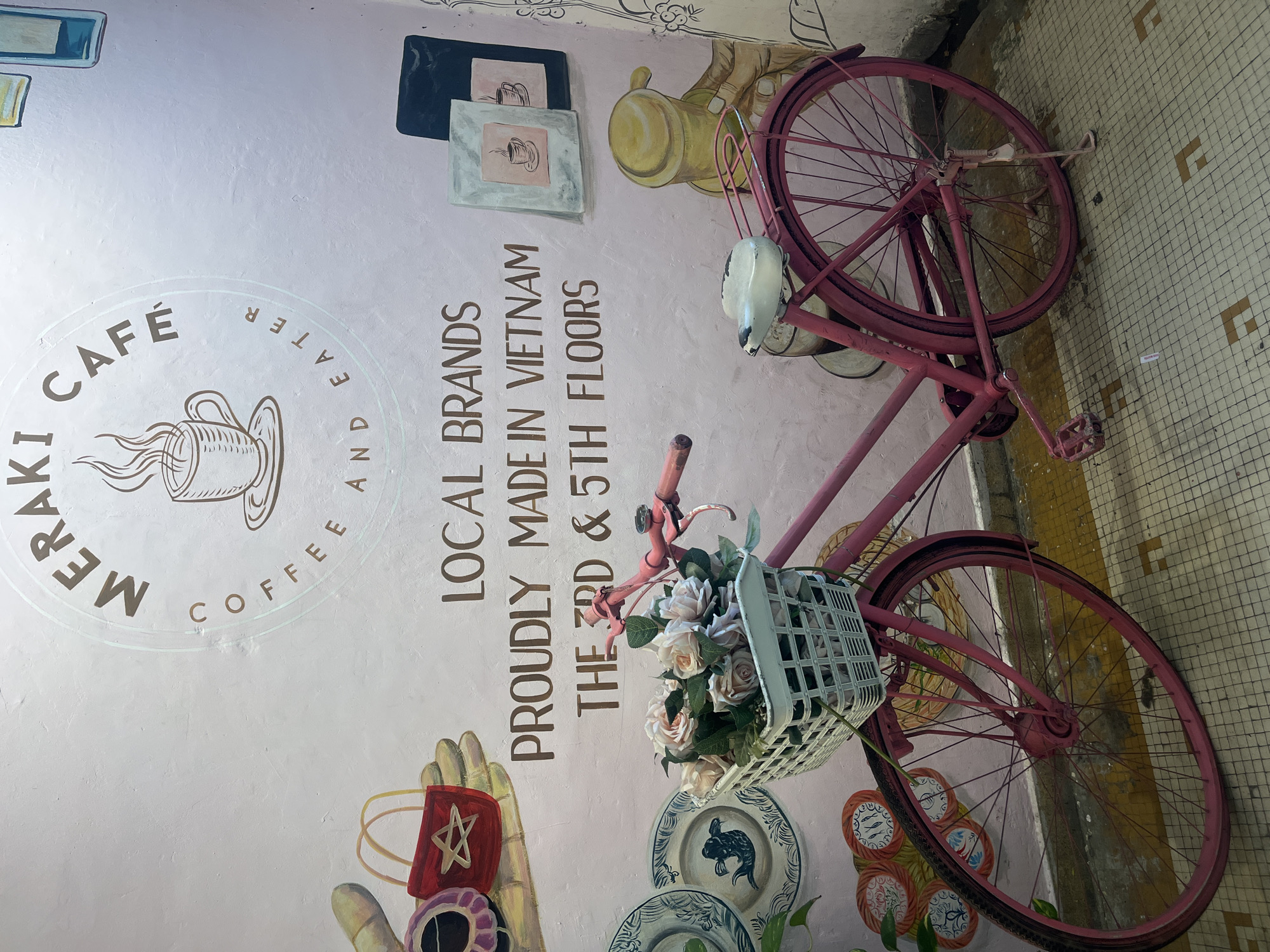 The image shows a pink bicycle with a basket of flowers parked in front of a pink wall. The wall is decorated with a mural that includes the words "MERAKI CAFE COFFEE AND EATER", "LOCAL BRANDS PROUDLY MADE IN VIETNAM THE 3RD & 5TH FLOORS", and drawings of coffee cups and packaging. The bicycle is the primary subject in the foreground. The background is a wall with artwork. The floor is tiled with a geometric pattern. The overall scene appears to be an outdoor or semi-outdoor setting, likely a cafe or shop. The image does not contain any discernible elements that specifically place it within Ho Chi Minh City, Vietnam, beyond the text on the wall indicating it is "PROUDLY MADE IN VIETNAM". There are no people or animals visible in the image. The lighting suggests it might be daytime or an indoor setting with artificial lighting.