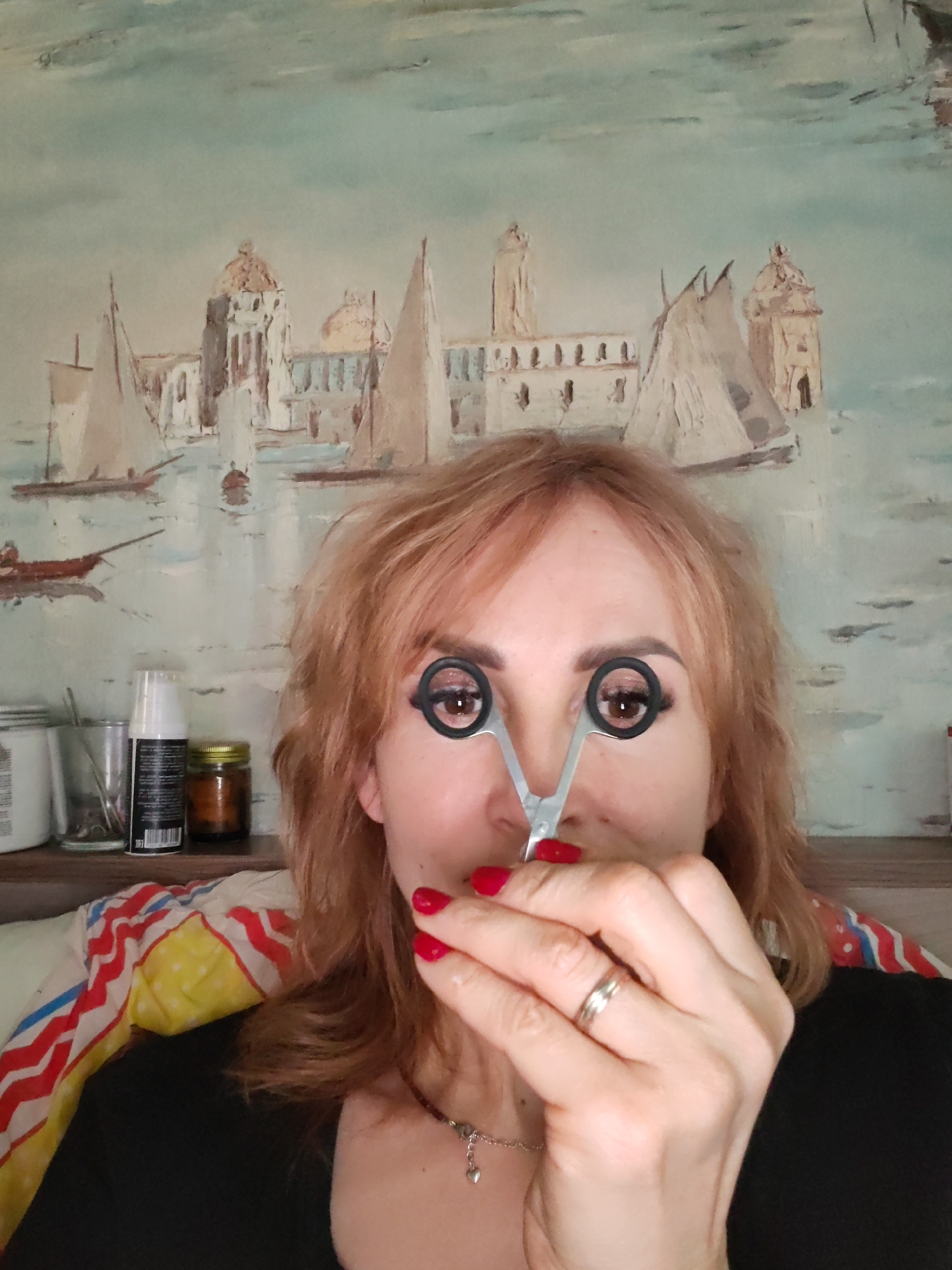 A woman is captured in a close-up shot, positioned indoors. She has reddish-blonde, somewhat disheveled hair and is wearing a black top. Her left hand, adorned with a silver ring on the ring finger and red nail polish, is holding a small silver pair of scissors. Playfully, she holds the open scissors up to her face, aligning her dark brown eyes with the black finger holes. A silver necklace with a small heart pendant is visible around her neck.
Behind her, an eye-level shelf holds various items: a white container with partially visible text "354003" and "HAIR", a clear glass with tools or brushes, a black spray can showing a barcode and "500 ml" and "MADE IN EU", and a small amber-colored jar.
The background is dominated by a wall hanging or painting depicting an old-world cityscape, possibly Venice or a similar European port. The artwork features numerous sailing boats with triangular sails on a body of water, alongside buildings with domes, towers, and arched windows, all rendered in muted blues, greens, and ochre tones. A colorful striped fabric, with red, yellow, and blue patterns, is visible over her left shoulder, possibly a blanket or scarf. The overall impression is one of a lighthearted moment in a personal, artistic setting in Chaikovsky, Russia.