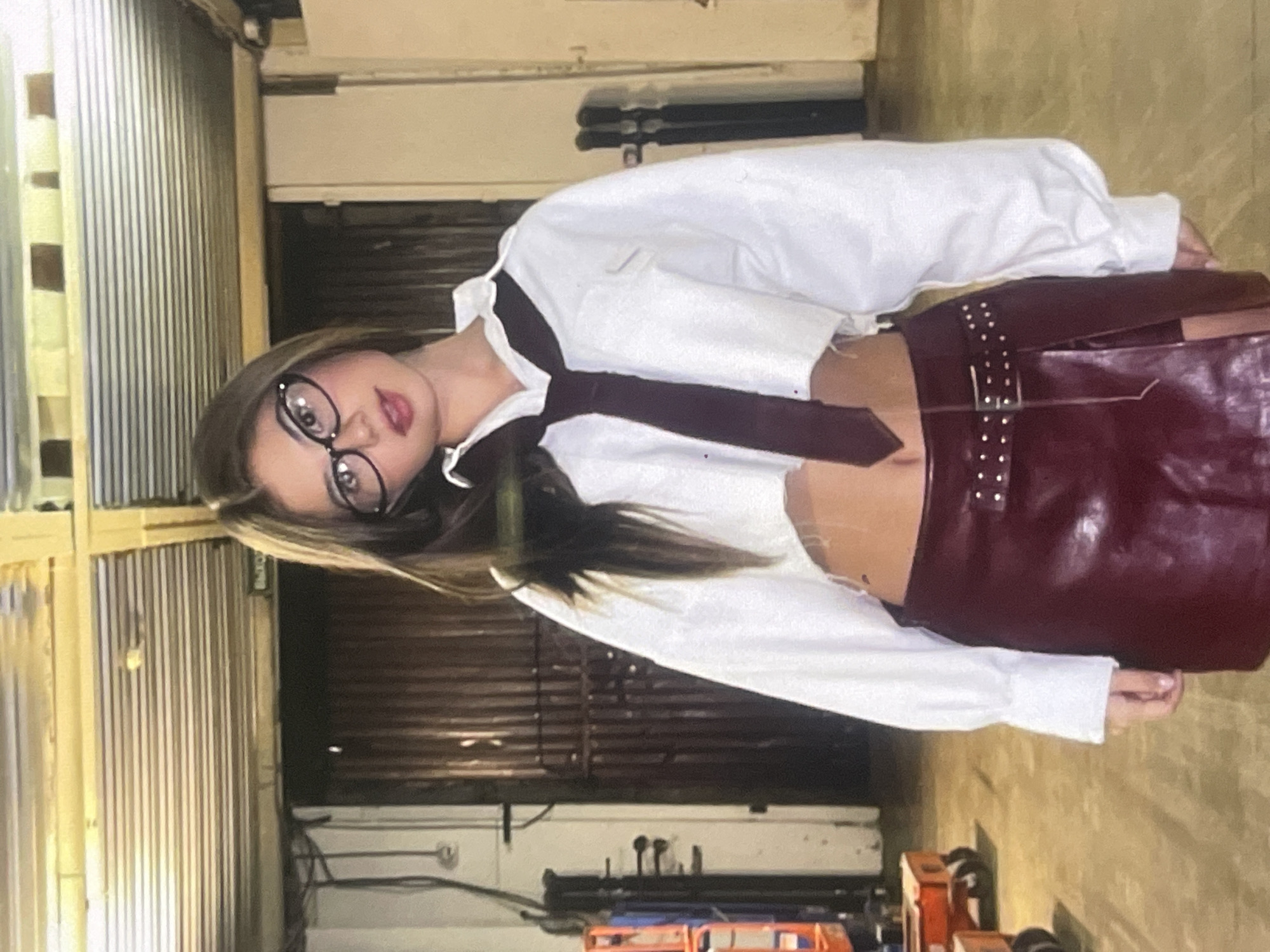 The image displays a young woman posing indoors. She has long, straight brown hair and is wearing round-framed glasses. Her attire consists of a white cropped long-sleeve shirt, exposing her midriff, paired with a dark tie. She also wears a dark red or burgundy leather-like skirt that features a studded belt. The woman looks directly towards the camera with a neutral expression.
The setting appears to be an industrial or storage-like environment. The background features a tall corrugated metal wall or door, a concrete floor, and various pieces of dark machinery or equipment, along with some visible cables. The lighting suggests an artificial indoor source. The photograph is taken at a slight angle. No discernible text is visible within the image. Based on the provided context, the location is Poltava, Ukraine. The time of day and external weather conditions are not evident from the indoor scene.