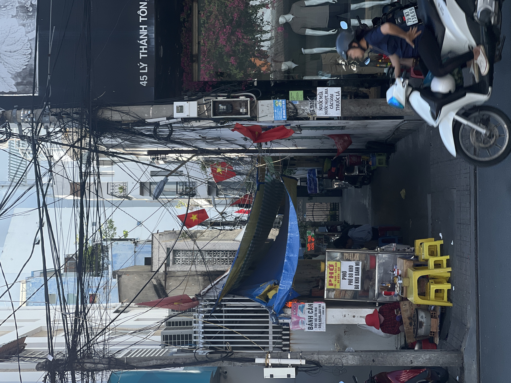 Here is a detailed description of the image:
**Location:** The image depicts a narrow street scene in Nha Trang Ward, Vietnam.
**Environment:** The street is flanked by buildings of varying heights and architectural styles. Overhead, a dense network of tangled electrical wires dominates the skyline. Several Vietnamese flags are strung across the street, adding color to the scene.
**People:** One woman wearing a red hat is seen sitting on the ground near a food stall, presumably preparing or serving food. Another person is riding a scooter down the street, partially visible.
**Objects:** A food stall selling "Banh Can" (Vietnamese mini pancake) and "Pho" (Vietnamese noodle soup) is positioned on the side of the street, with yellow plastic stools nearby. The shop has signage in Vietnamese. Other notable objects include motorbikes parked along the street, various signs and advertisements, and a partially visible blue tarp hanging above the street.
**Actions:** The woman is engaged in activity related to her food stall. The scooter rider is navigating the street.
**Interactions:** There is a potential customer relationship between the woman and the potential customers.
**Additional Details:**
- The sign for "45 Ly Thanh Ton" can be seen at the top of the image.
- A white and blue scooter is seen in the street.