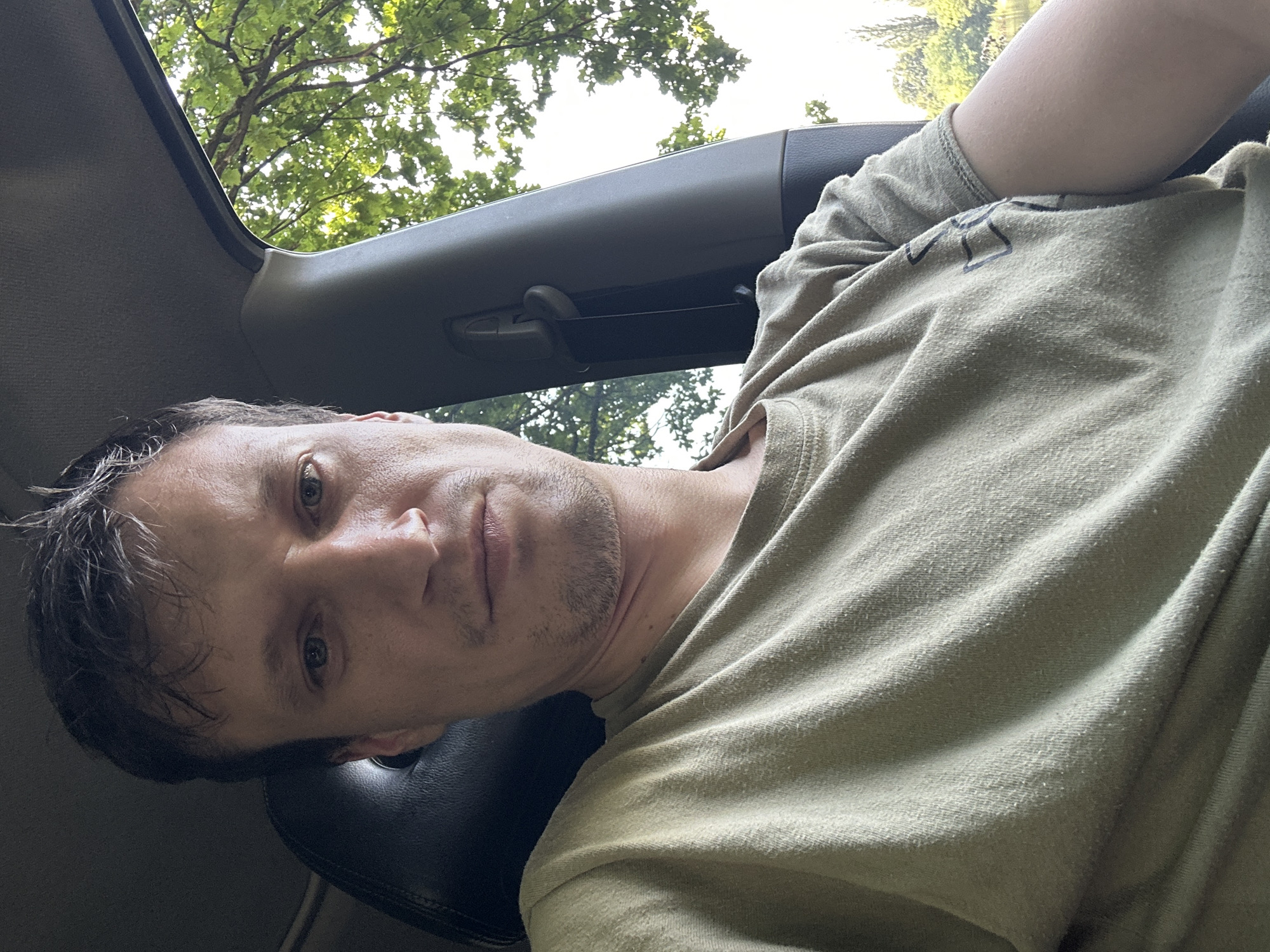 A man is sitting in the driver's seat of a vehicle. He is wearing a light green t-shirt. The man is looking at the camera. The car interior is visible, including the dashboard and part of the passenger seat. Through the open window, trees and foliage are visible, suggesting an outdoor location. The background hints at a sunny day with natural light filtering through the leaves. The man's expression is neutral. The photo appears to be a selfie taken inside the car. The location is not explicitly stated to be in Makiv, Ukraine.