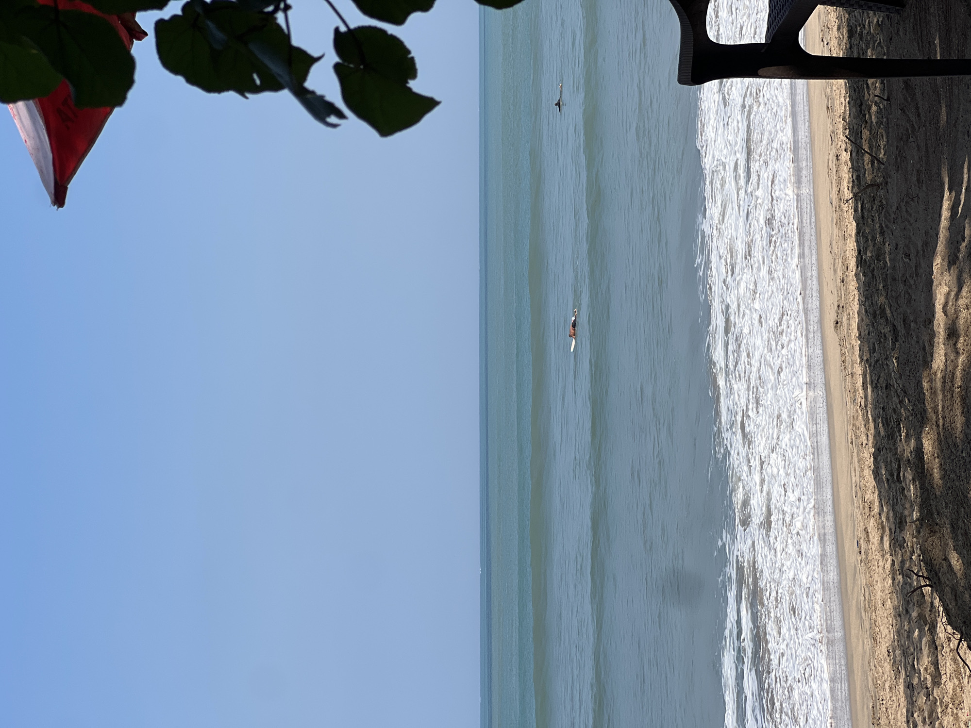 In Kuta, Indonesia, a clear sky presides over a beach scene. The ocean, with its turquoise and deep blue hues, features gentle waves lapping onto the sandy shore. Two individuals are visible in the water, both lying on surfboards. One person is closer to the shore, while the other is further out. The foreground of the image is dominated by sand, with shadows cast from an unseen object, and a dark plastic chair is positioned on the right side of the frame. A portion of a red and white umbrella is visible in the upper right corner, partially obscured by lush green leaves of a tree. The overall atmosphere is one of serene beach activity under a bright, sunny sky.