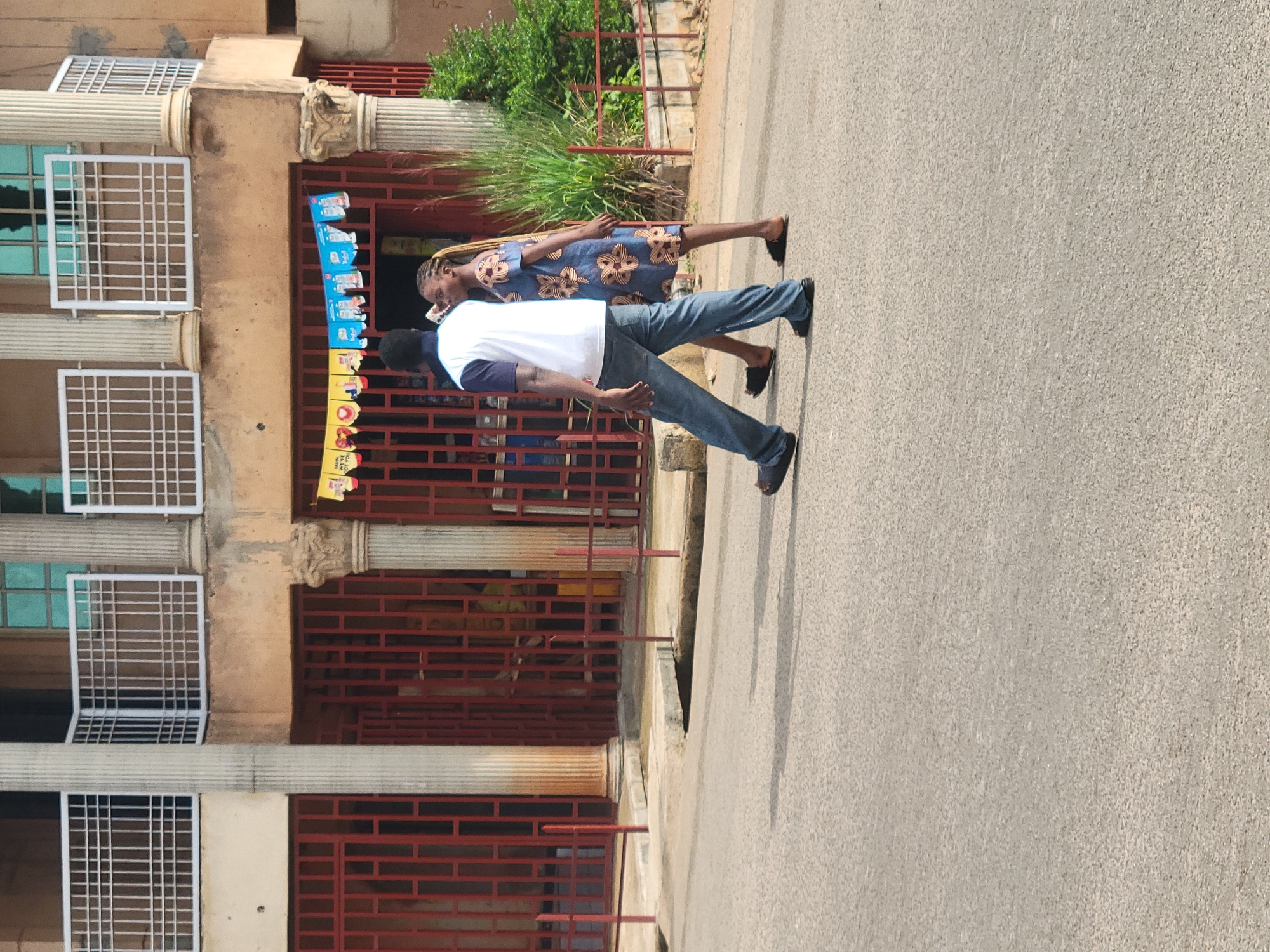 A man and a woman are walking on a paved street in front of a building. The man is wearing a white t-shirt with blue accents and blue jeans, and he is walking to the left of the frame. The woman is wearing a blue dress with a floral pattern and is walking to the right of the frame, slightly behind the man. Behind them, a two-story building with red metal gates and ornate columns is visible. A string of colorful banners hangs across the front of the building, and a green tree is to the right. The ground is a paved street with shadows cast by the people and objects.