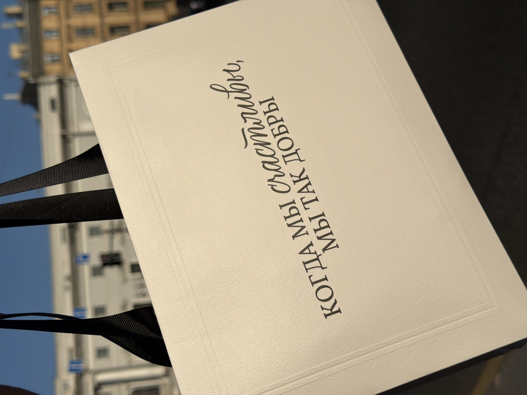 A close-up, eye-level shot shows a white gift bag with black ribbon handles held up against an outdoor background. The gift bag is textured and has embossed lines on its surface. Dark, elegant script is printed on the bag in Russian. The top line reads "КОГДА МЫ" and the second line reads "МЫ ТАК ДОБРЫ" with a cursive word "счастливы," between them. In the blurred background, a light-colored building with multiple windows and some architectural details is visible, along with a traffic light and what appears to be a segment of a yellow building. The overall impression is that of a bright, sunny day.
